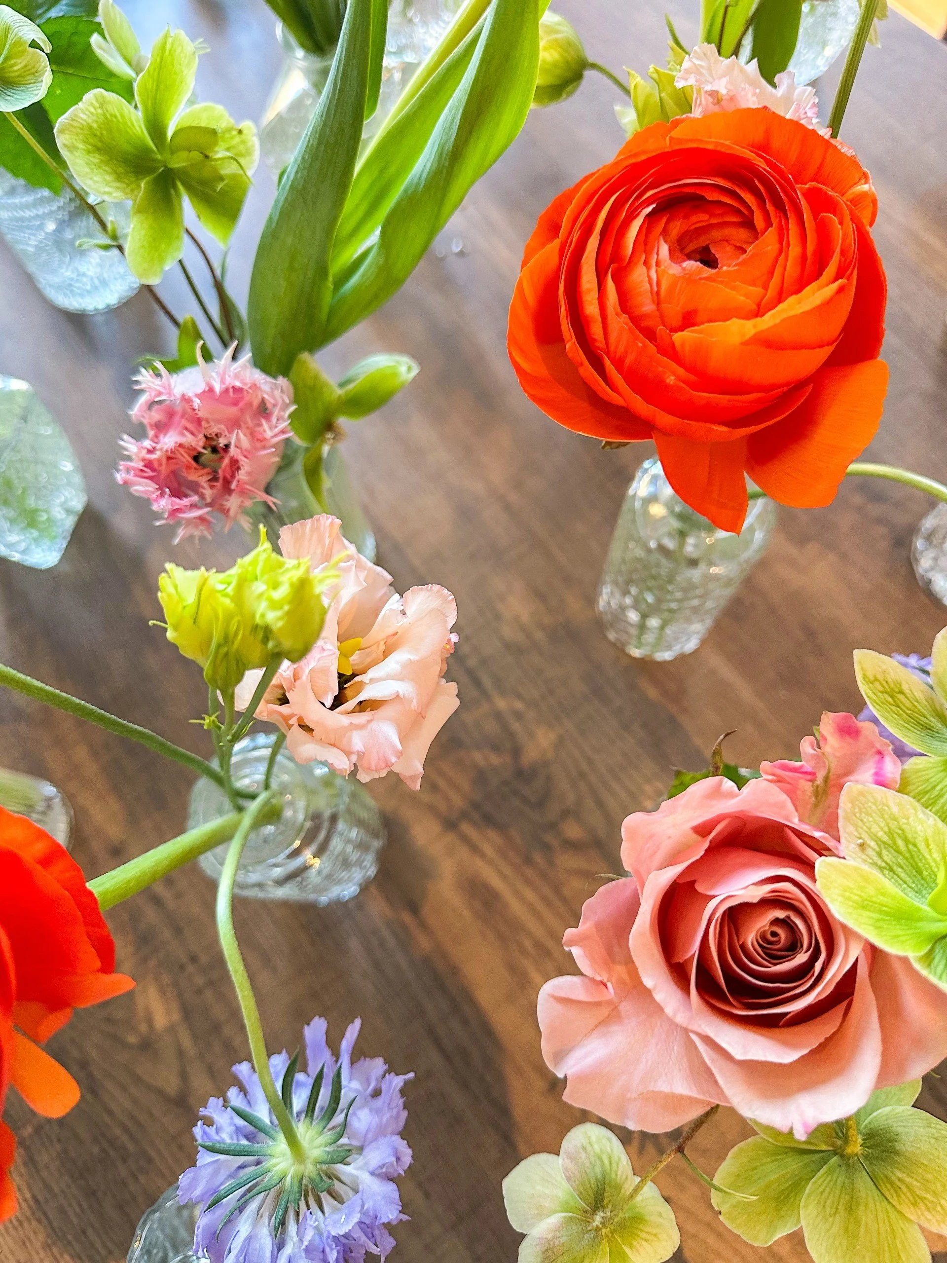 Various colorful flowers in small glass vases on a wooden table, including an orange ranunculus, a pink scabiosa, a purple starflower, and a green hydrangea.