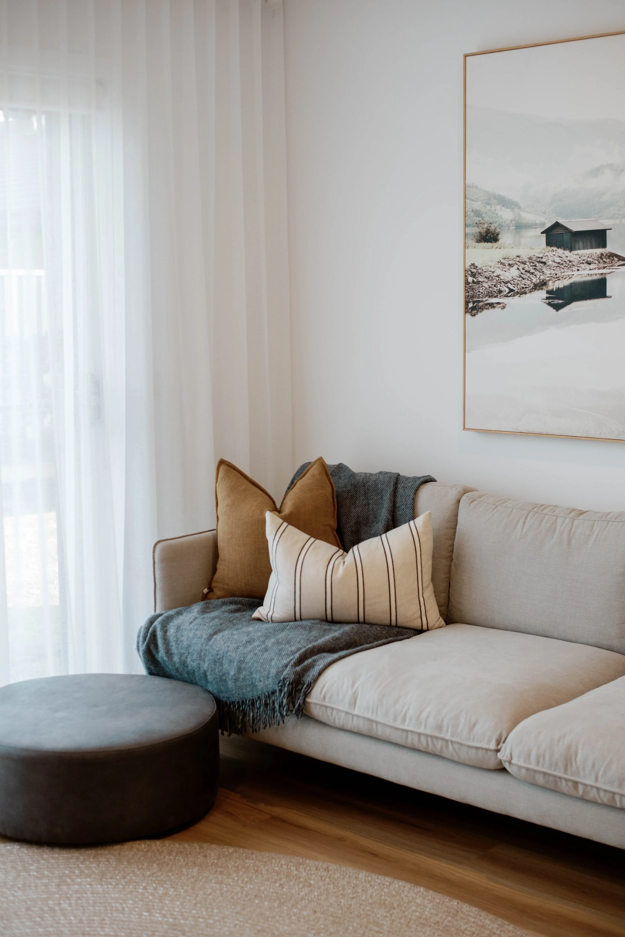 Display home for a builder in Bendigo, Victoria. Living room with beige sectional sofa, brown and striped pillows, dark round ottoman, light-colored curtains, framed landscape wall art, wooden floor, and beige carpet.