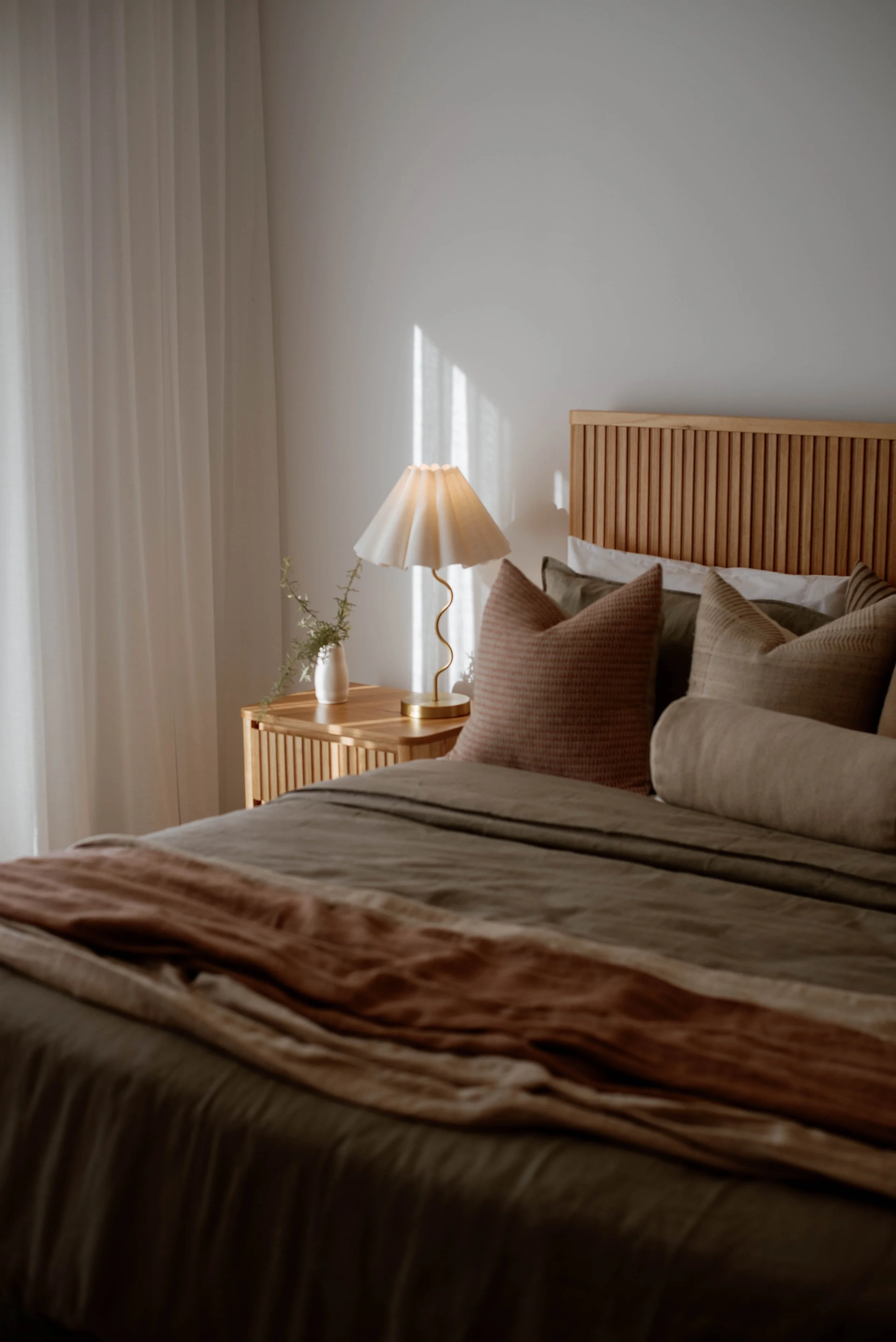 Display home for a builder in Bendigo, Victoria. A bedroom with a wooden headboard, beige pillows, a bedside table with a lamp, a white vase with green foliage, and white curtains letting in natural light.