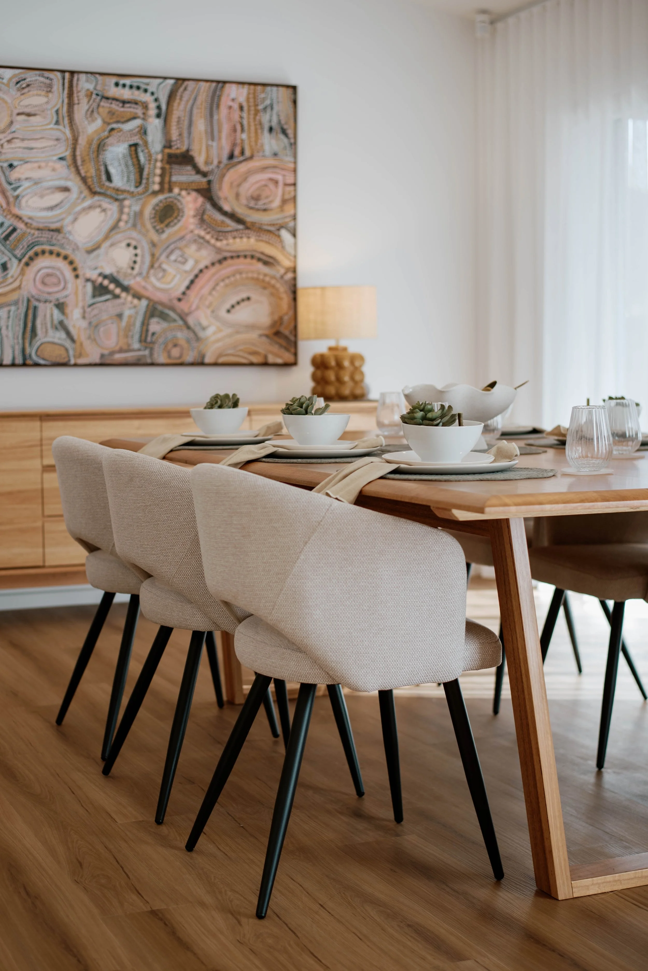 Display home for a builder in Bendigo, Victoria. A dining table set for four with white bowls, napkins, glasses, and small succulents in white bowls. A large abstract painting hangs on the wall behind, and a wooden sideboard with a lamp. 