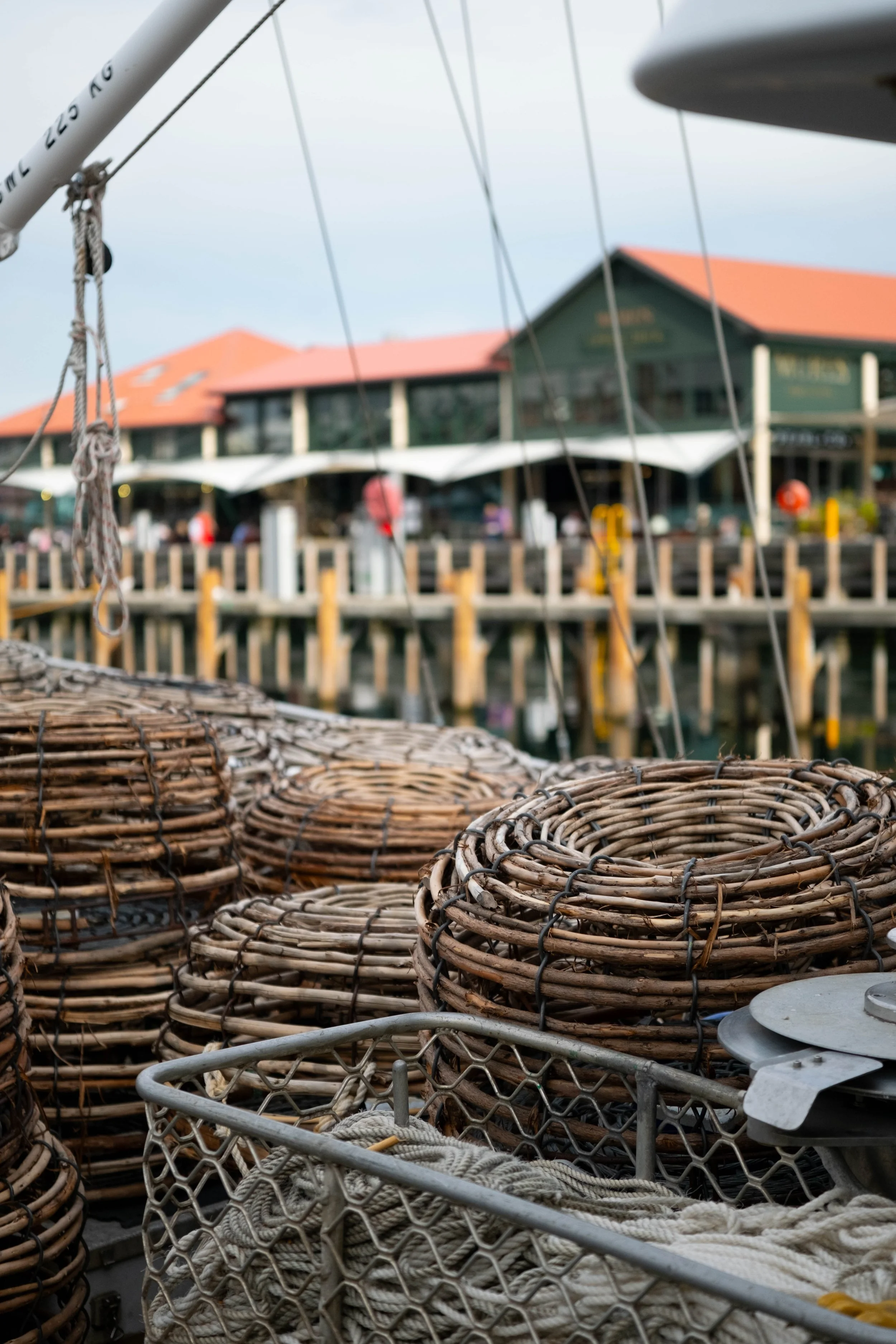 Close-up of woven wicker lobster traps with metal rings in the foreground, with a blurry background of a pier, restaurant, and people by the water.