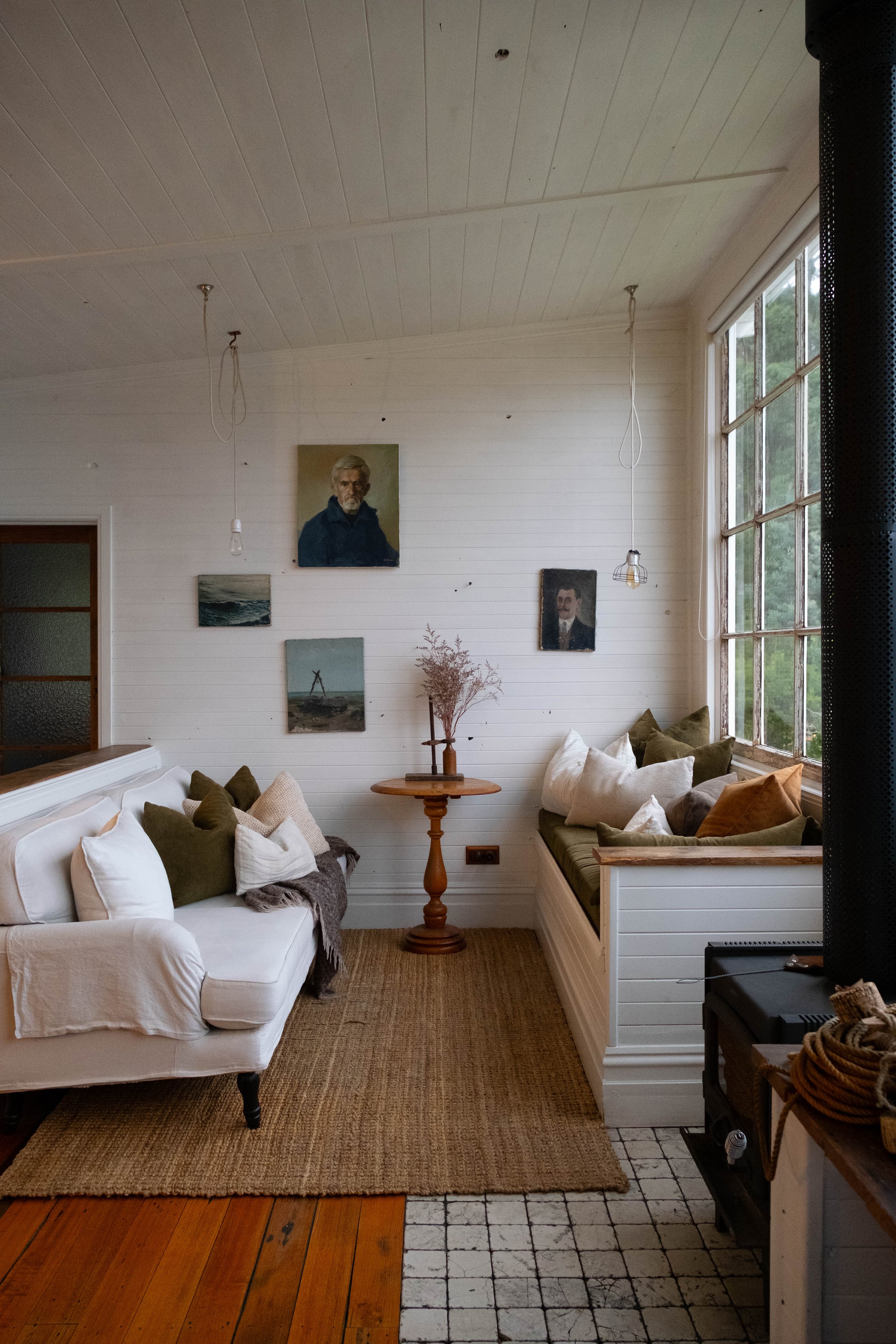 A cozy living room features white walls decorated with paintings, a white sofa, a wooden side table with dried flowers, and a corner built-in bench with pillows by a large window. Several hanging light fixtures are visible, and the room has a warm, i