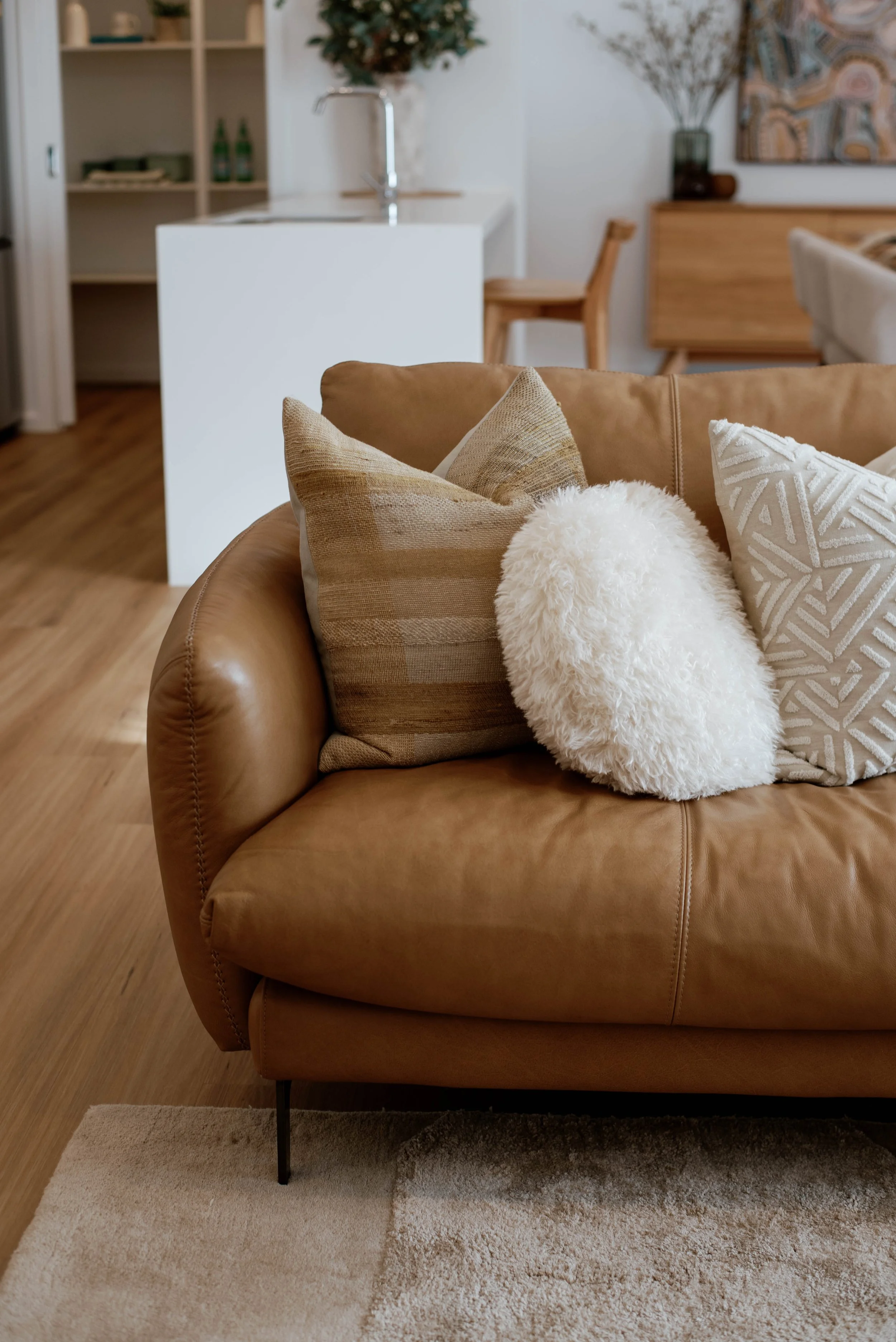 Display home for a builder in Bendigo, Victoria. Living room with a tan leather sofa, decorative pillows, a patterned area rug, a wooden dining table, and a kitchen area in the background.