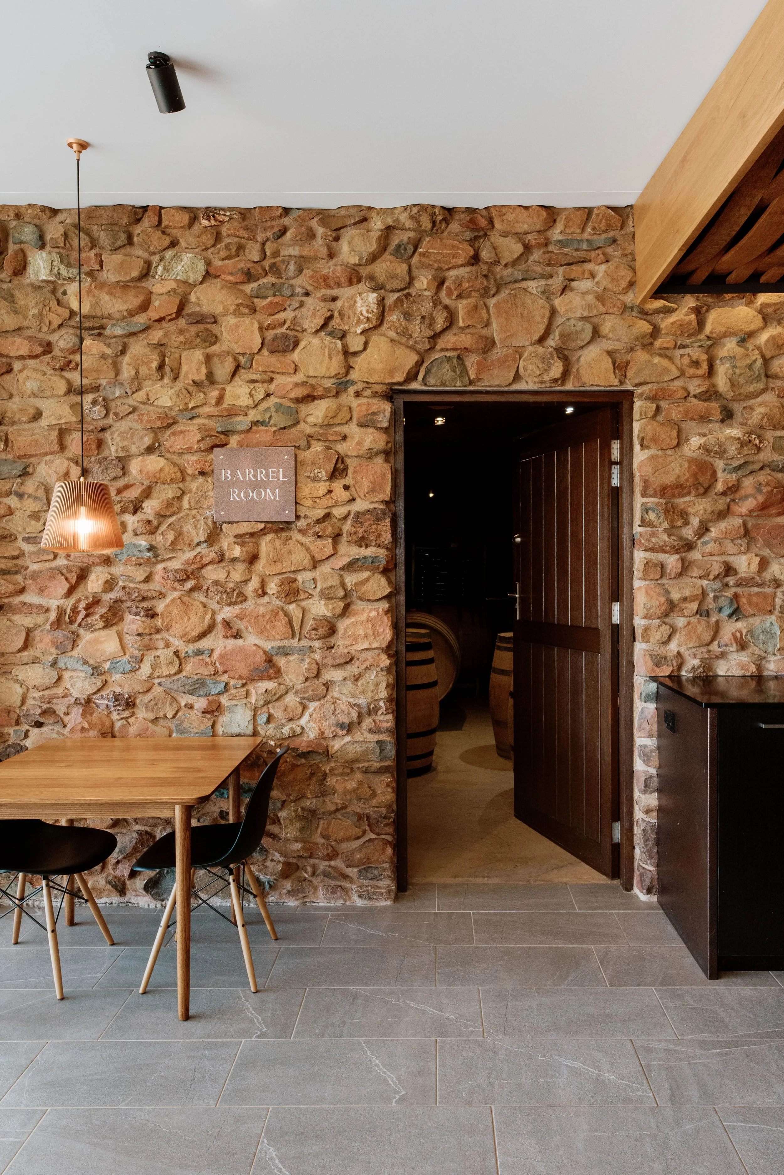 Scandinavian inspired winery in Heathcote, Victoria. Interior of a room with a stone wall, wooden furniture, a hanging lamp, and a sign that reads 'Barrel Room.'