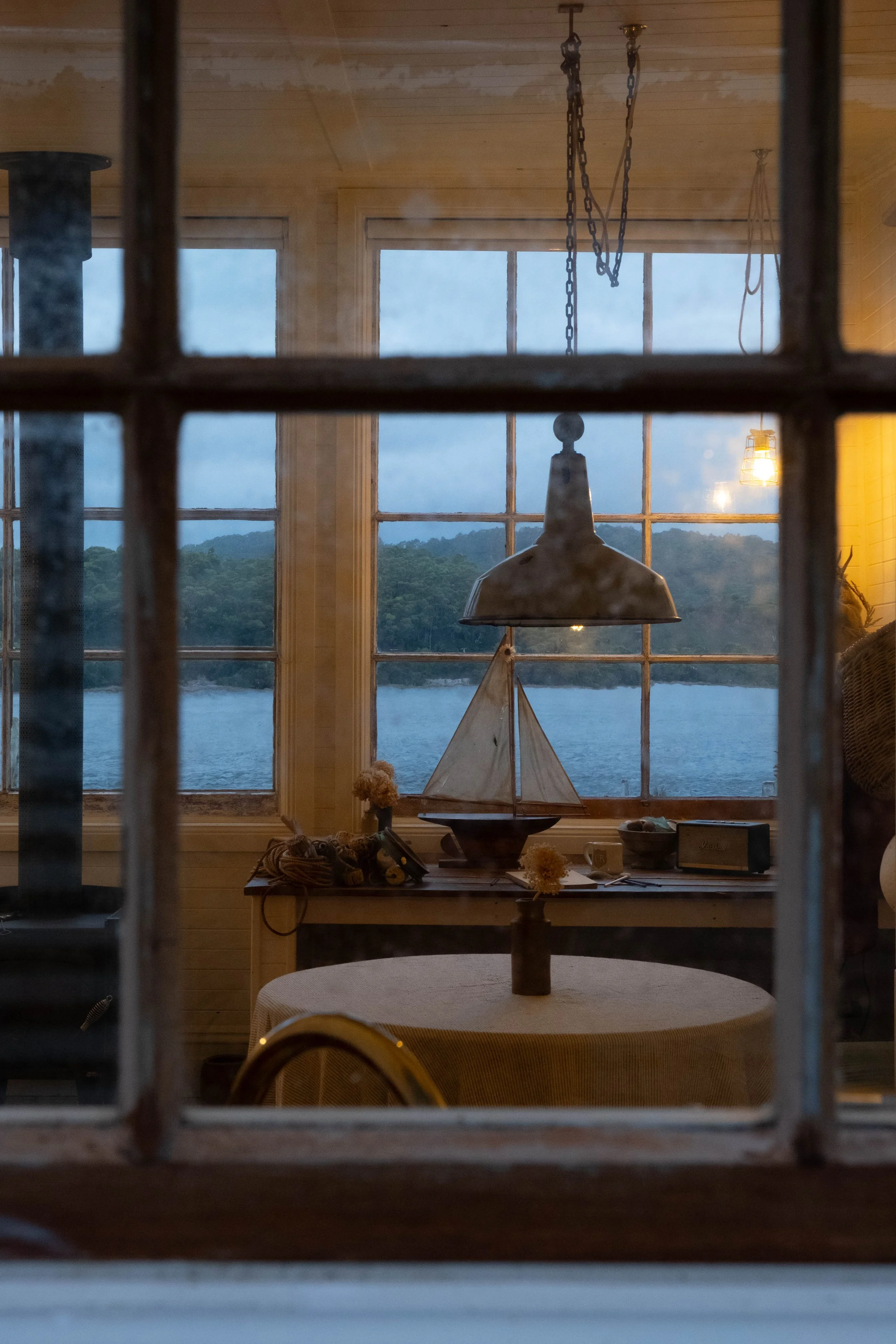 View through a window showing a room with a nautical theme. There is a small white sailboat model on a table, with a window behind showing a river and green hills in the distance. The room has wooden walls and a hanging lamp.