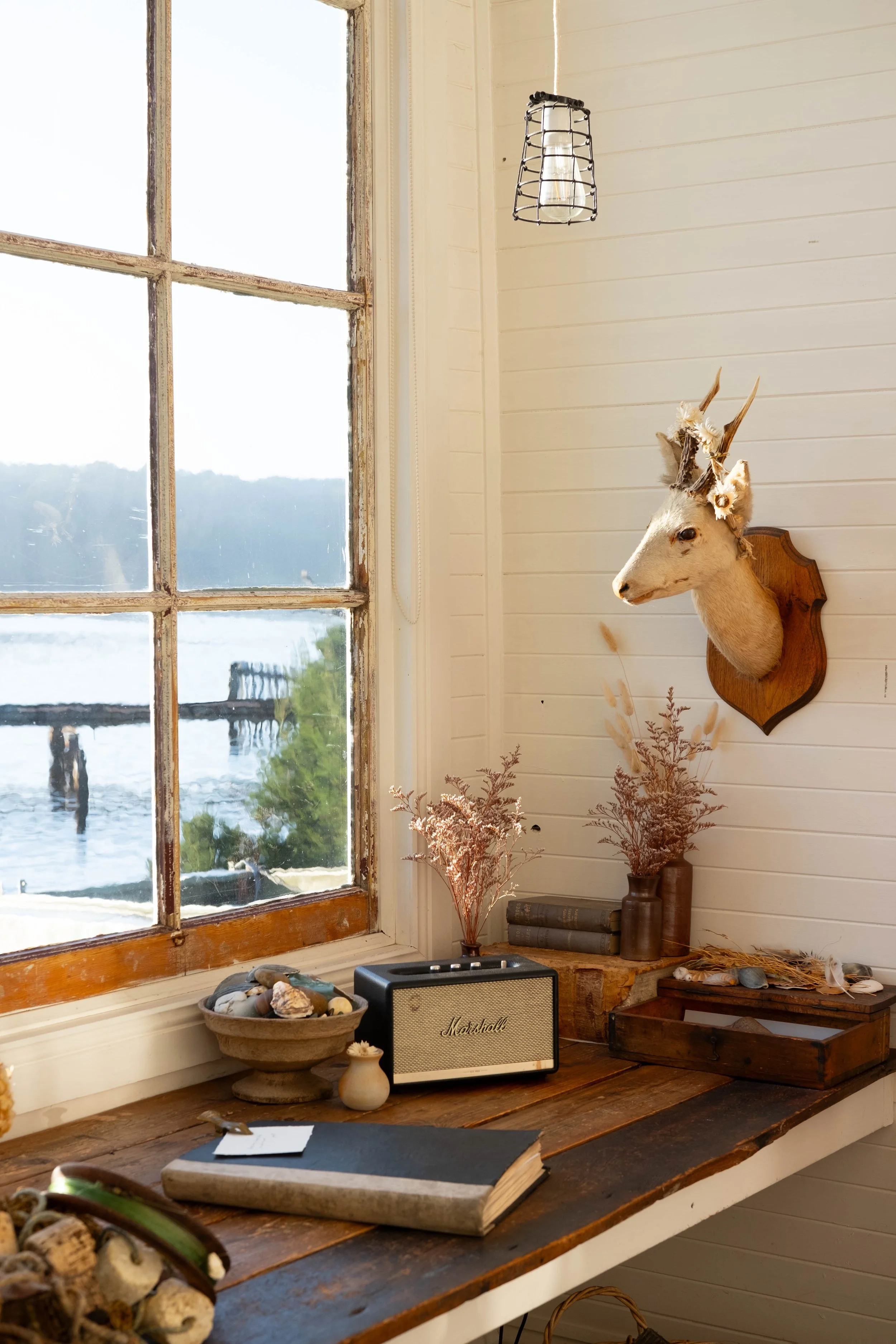 A rustic interior scene featuring a wooden table near a window with a view of water and a dock outside. On the table, there is a bowl of seashells, a small speaker, an open book, and some decorative items. Mounted on the wall is a taxidermy deer head