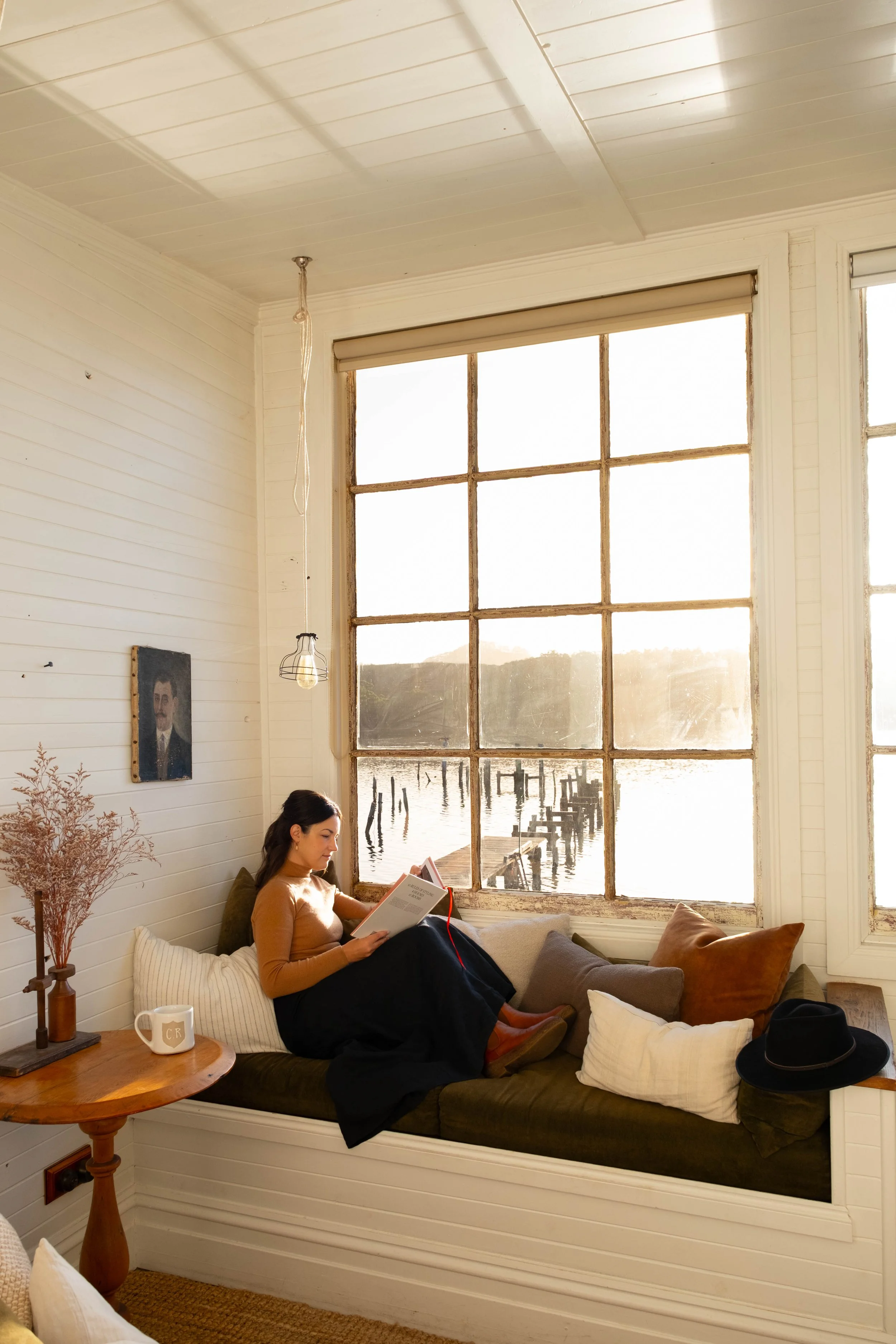 A woman sitting on a built-in window bench, reading a book, with a large window behind her showing a body of water and a dock outside. The room has white wooden walls and ceiling, with a small wooden side table holding a mug and a decorative vase on her left. There are multiple cushions on the window bench, and a hat and some pillows on the floor.