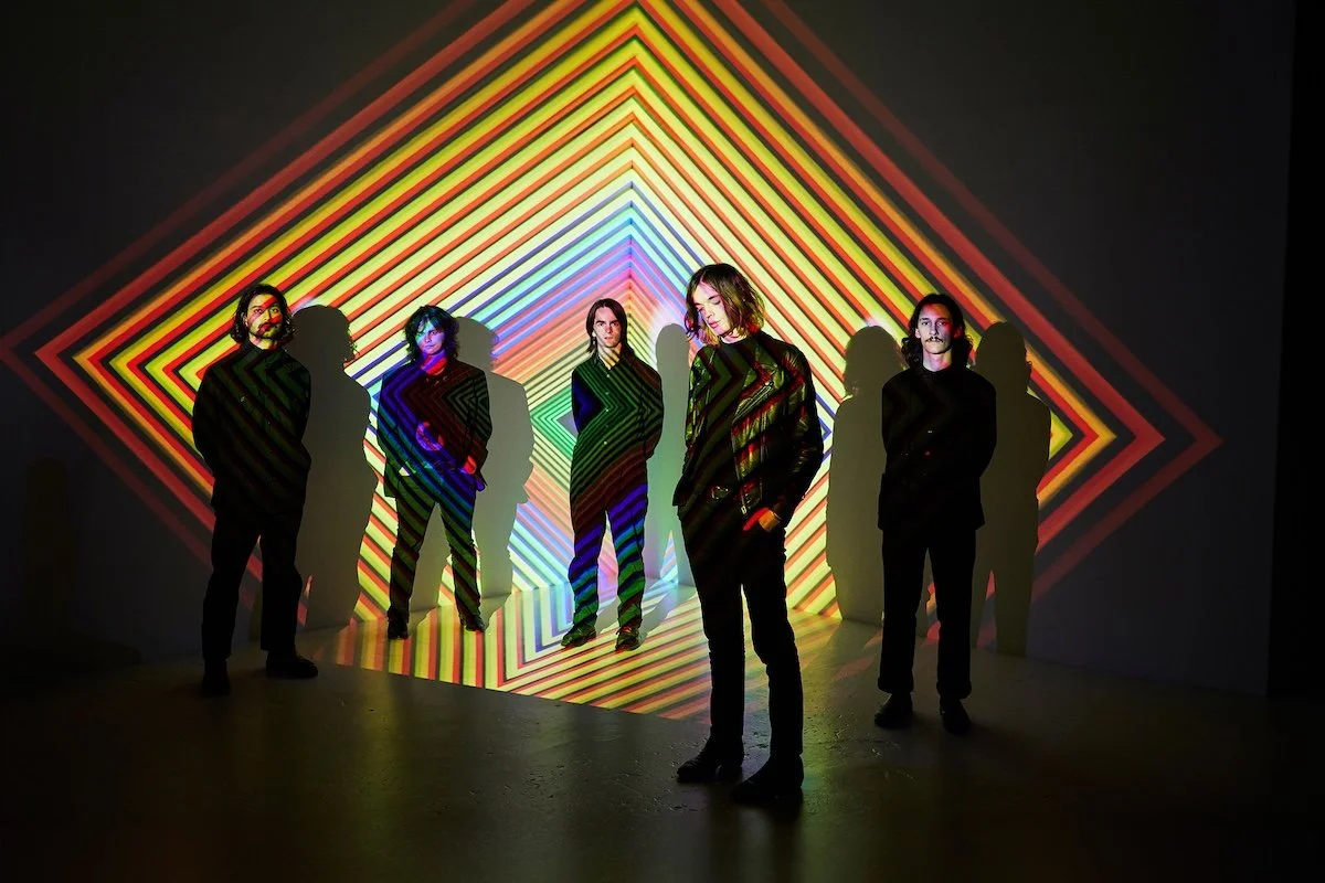 Psych-rock band Druid Fluids posing in front of a vibrant, multi-colored geometric light projection with rainbow diamond patterns and silhouettes.