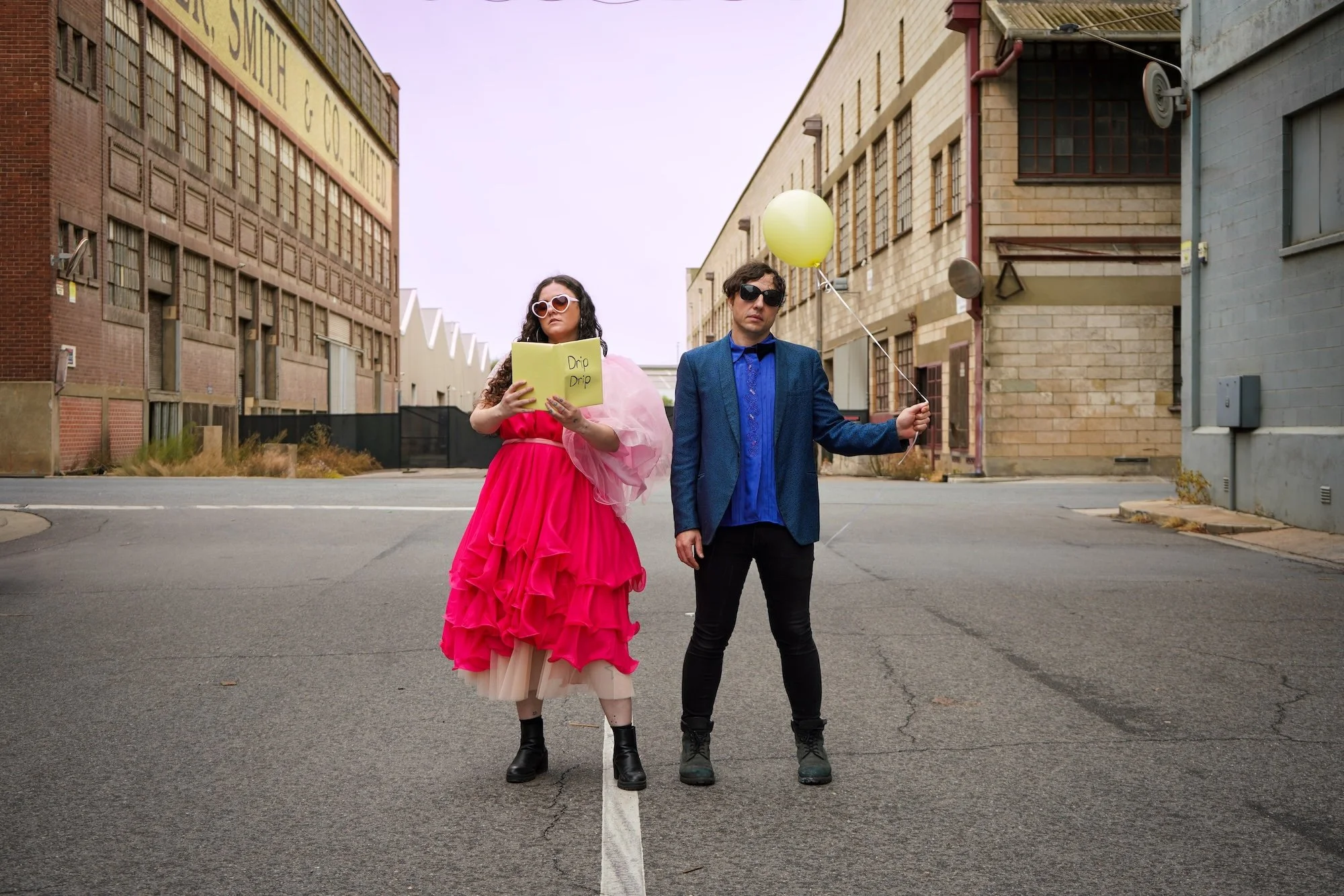 Miss Orange Juicy duo posing in an industrial street; one member in a bright pink ruffled dress and the other in a blue blazer holding a yellow balloon.