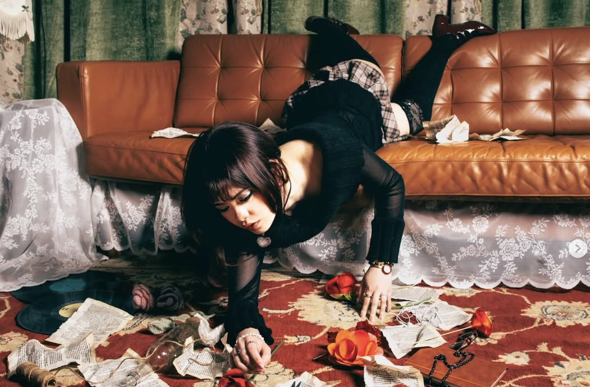 Indie artist Mariae Cassandra is sprawled on a floor covered in paper and records, with her legs draped over a brown leather couch.
