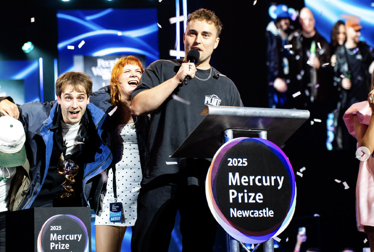 Sam Fender at the Mercury Prize with his bandmates. He speaks into a microphone at a podium with the 2025 Mercury Prize logo.