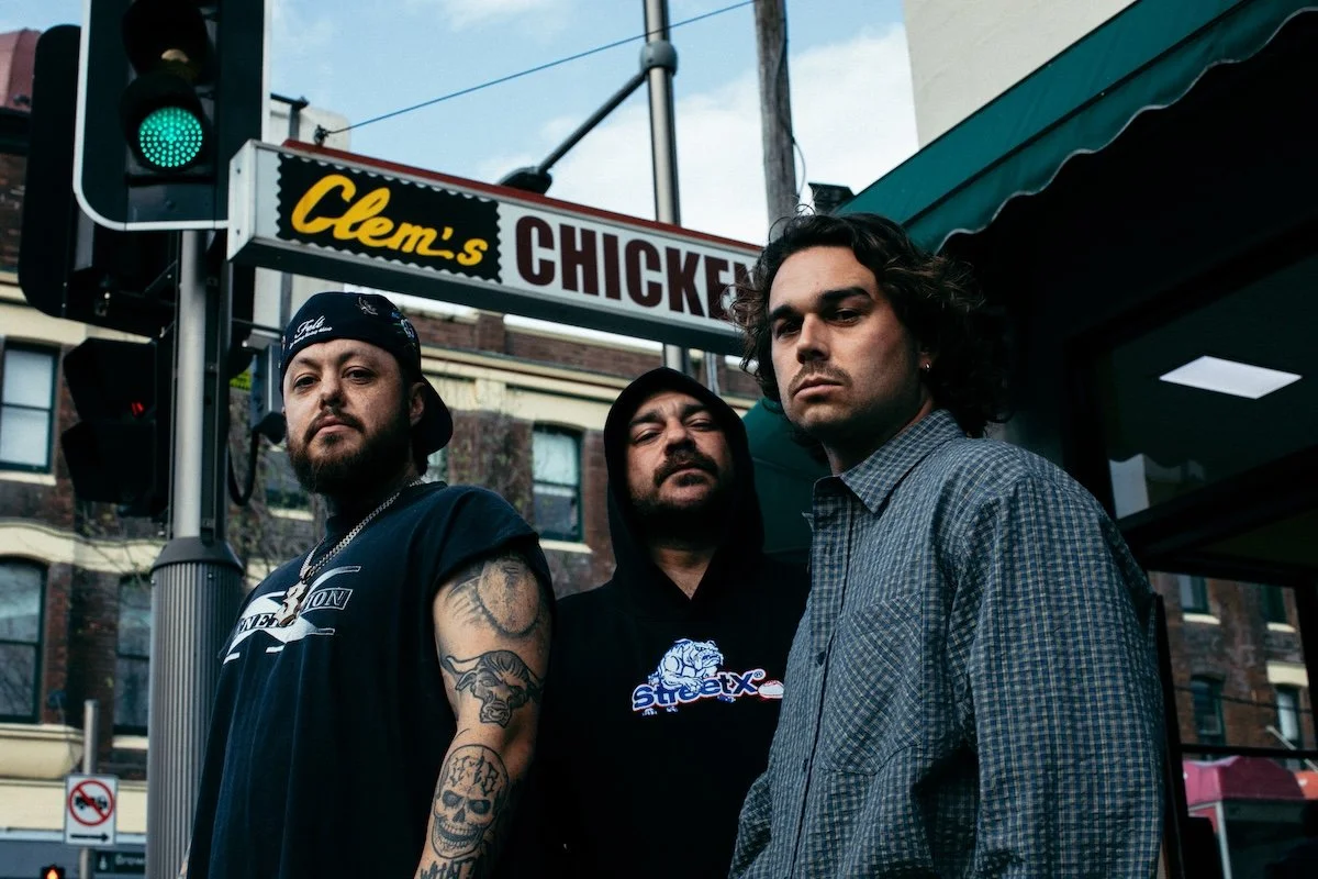 Australian hip-hop group 3% (Hazy, Trials, and Dallas Woods) standing together on a city street below a "Clem's Chicken" sign.