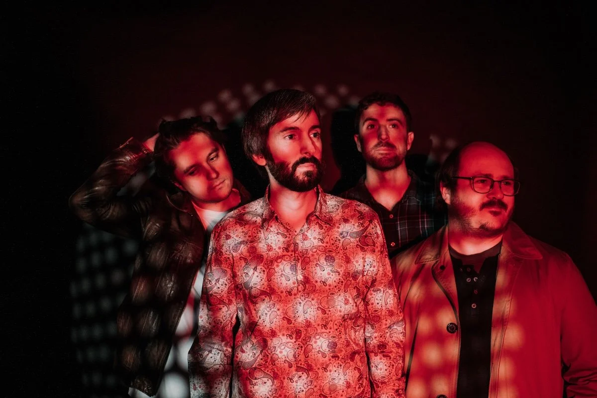 Promotional band photo of July Morning. The four members are bathed in moody red and white patterned lighting, featuring a lead singer in a paisley shirt.