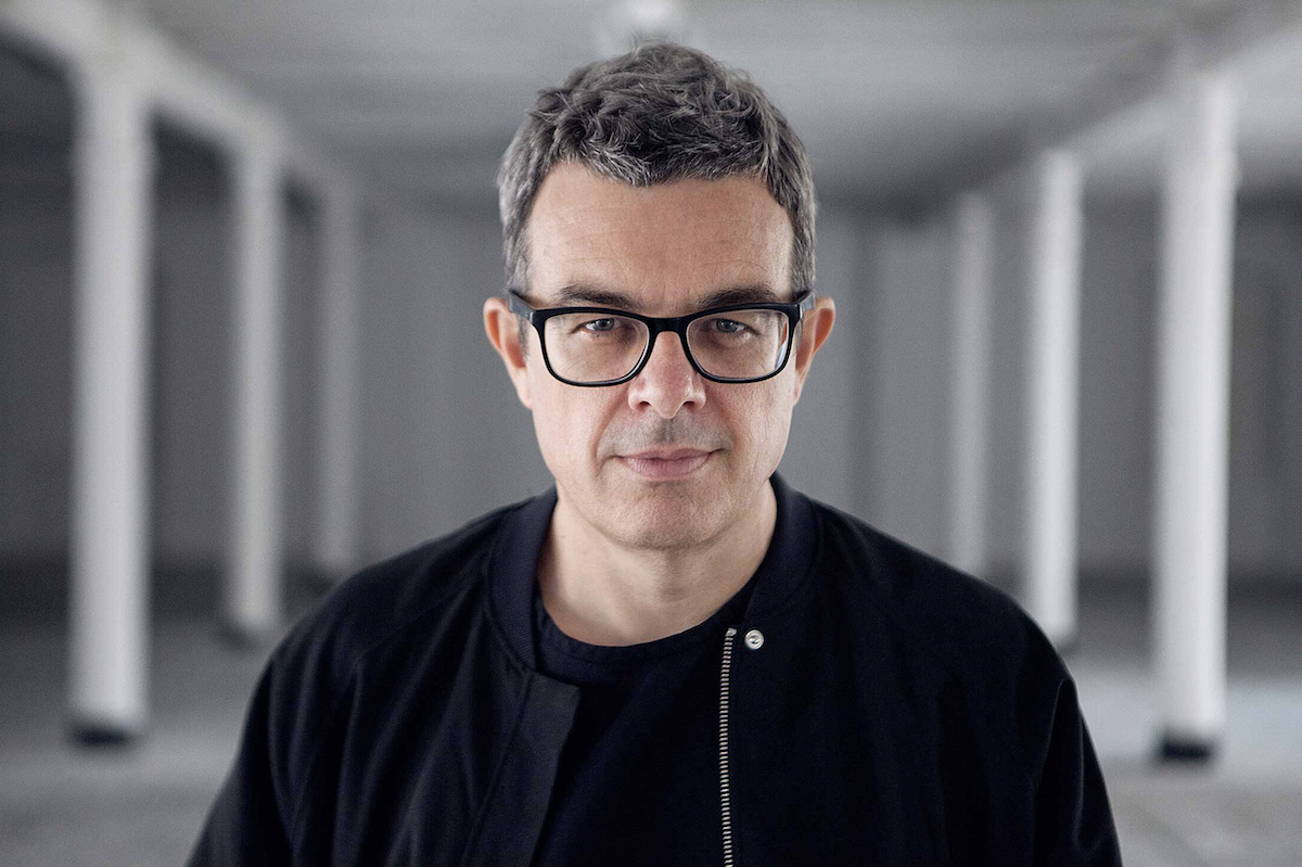 Portrait of Mat Schulz, co-founder of Unsound Festival, wearing glasses and a black jacket, looking forward against a blurred industrial background.