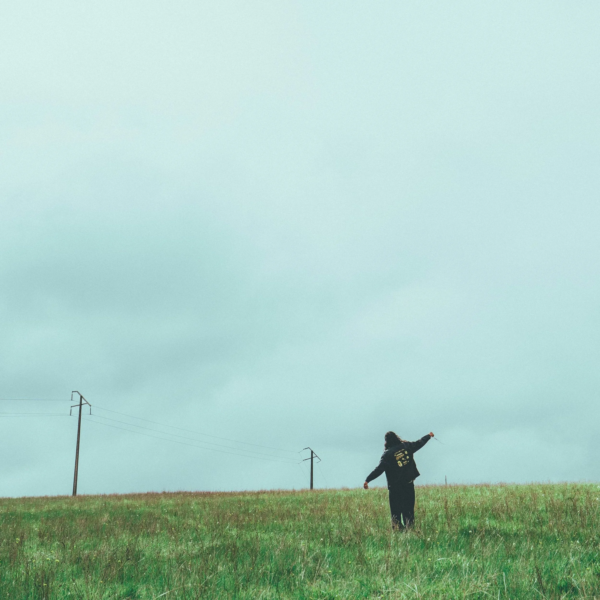 Single artwork for Spell by TUSHAR, featuring a distant figure in black standing in a vast green field with power lines under a massive cloudy sky.