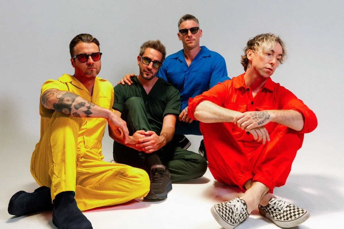 Pop-punk band All Time Low sitting in colorful jumpsuits: Alex Gaskarth, Jack Barakat, Zack Merrick, and Rian Dawson.