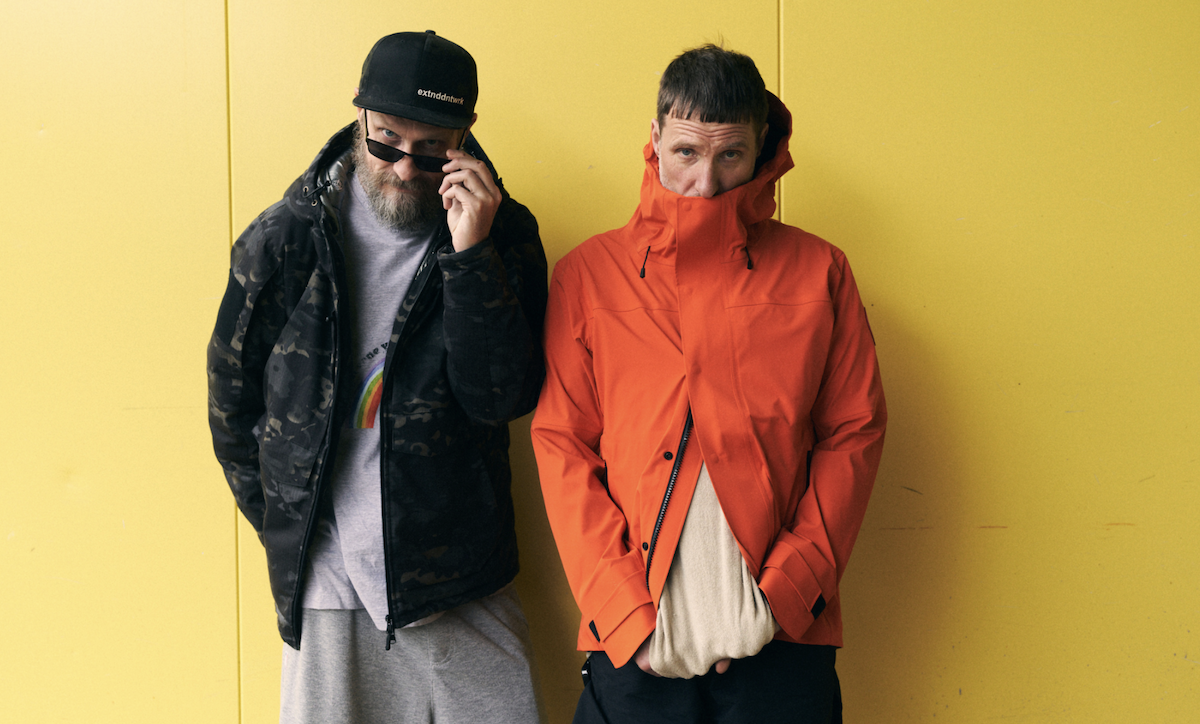 Sleaford Mods band members Jason Williamson and Andrew Fearn against a yellow wall in an orange jacket and a camo jacket.