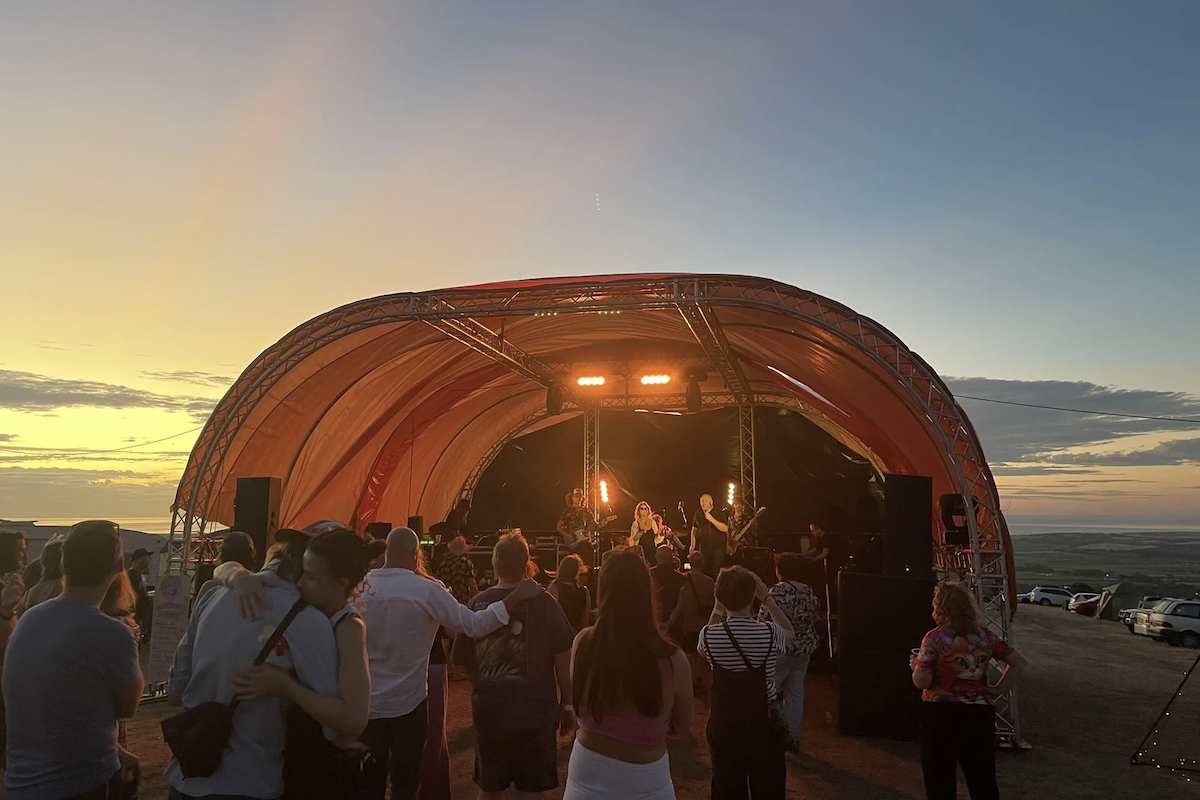 Concert at Dougstock music festival at sunset. A crowd faces a band performing on an outdoor stage with an orange arched canopy.
