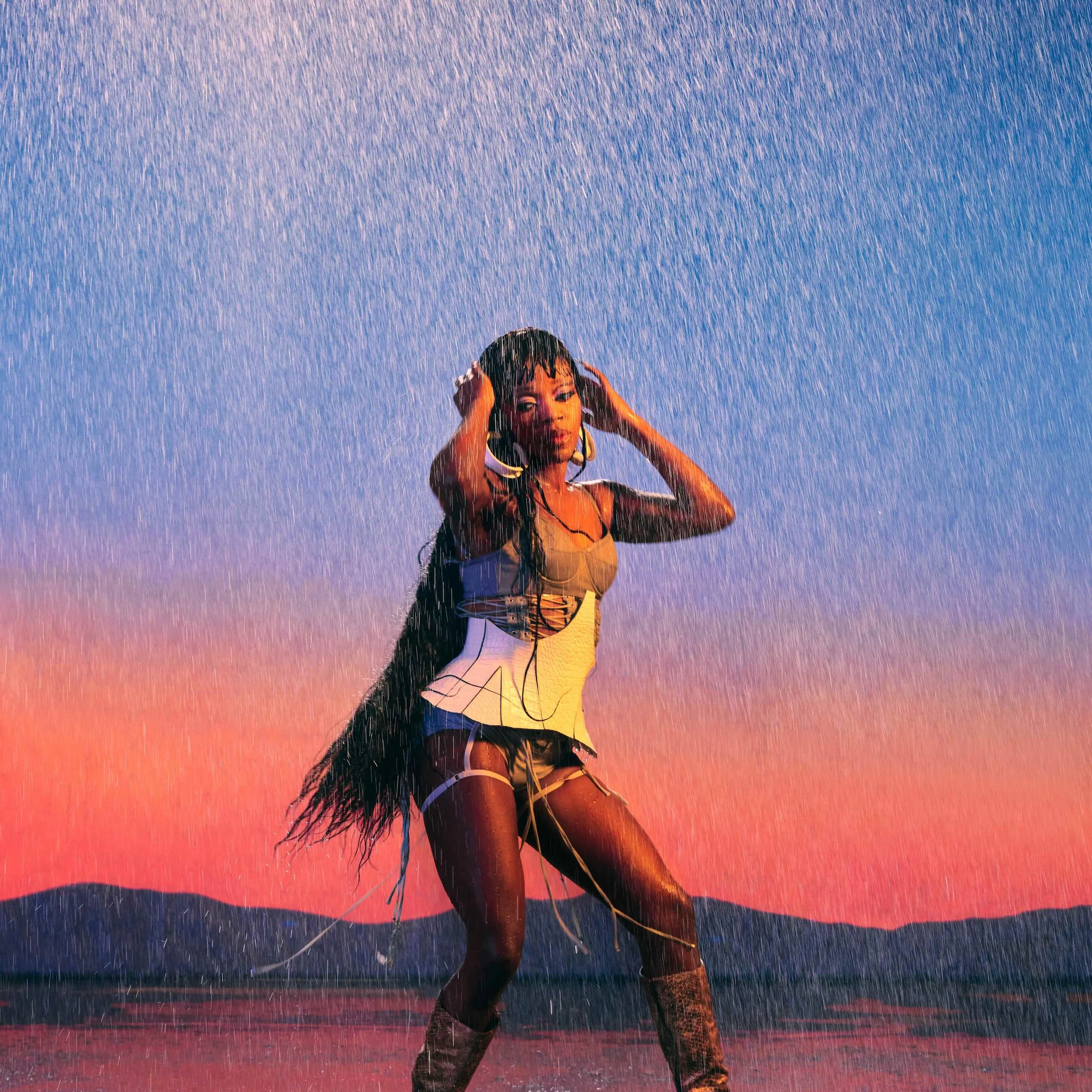 Tkay Maidza poses in falling rain against a vibrant sunset for the 'Pressed' single artwork, wearing a white corset and boots in a desert landscape.