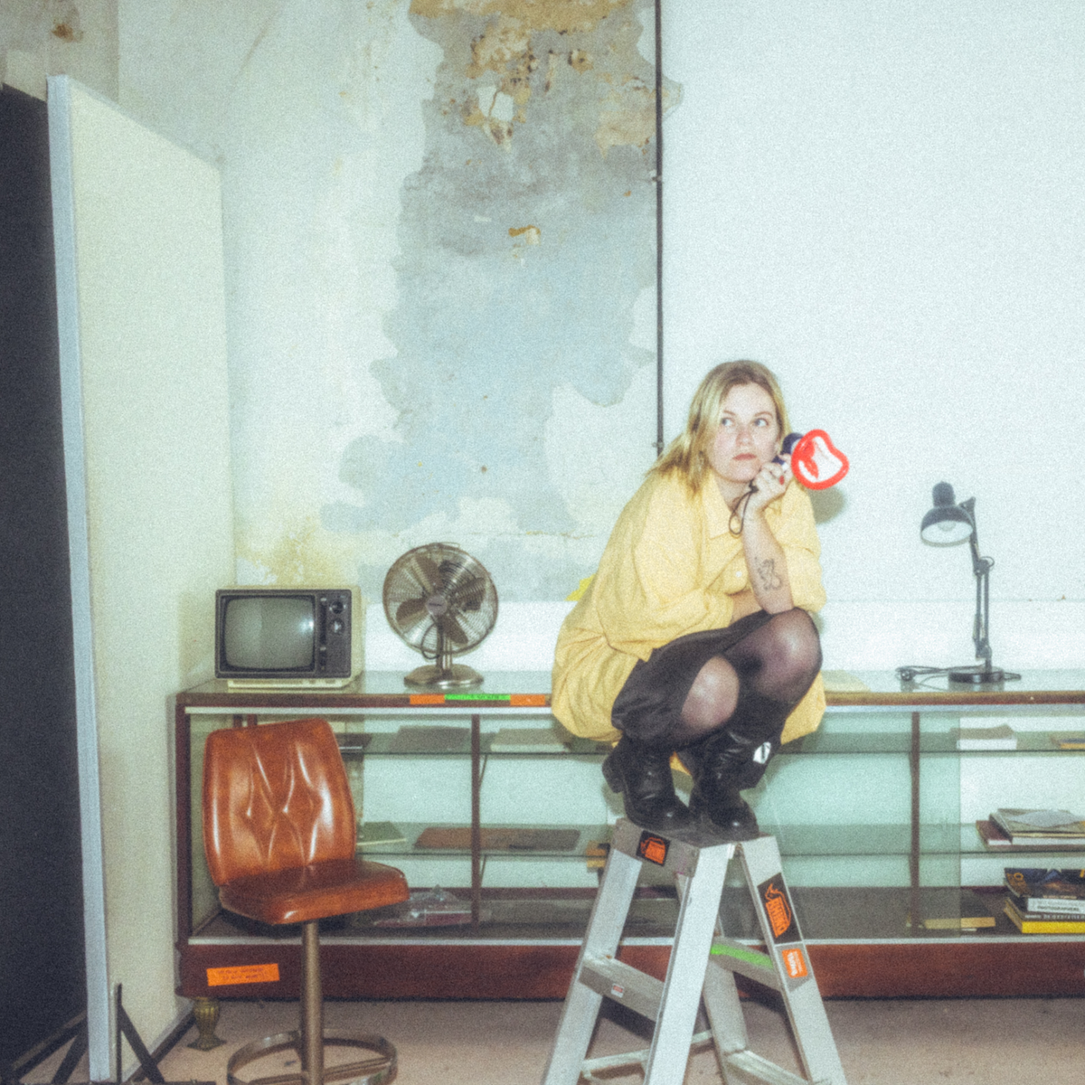 Album cover for Romanie's "It's Not That Funny" featuring the artist crouching on a stepladder in a room with vintage furniture and a weathered wall.