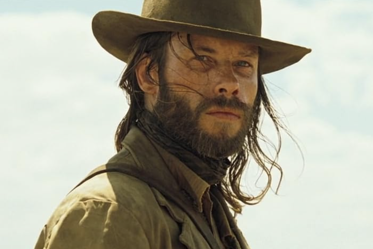 Guy Pearce as Charlie Burns in the 2005 Australian Western film The Proposition. He wears a dusty brown hat and coat against a bright outback sky.