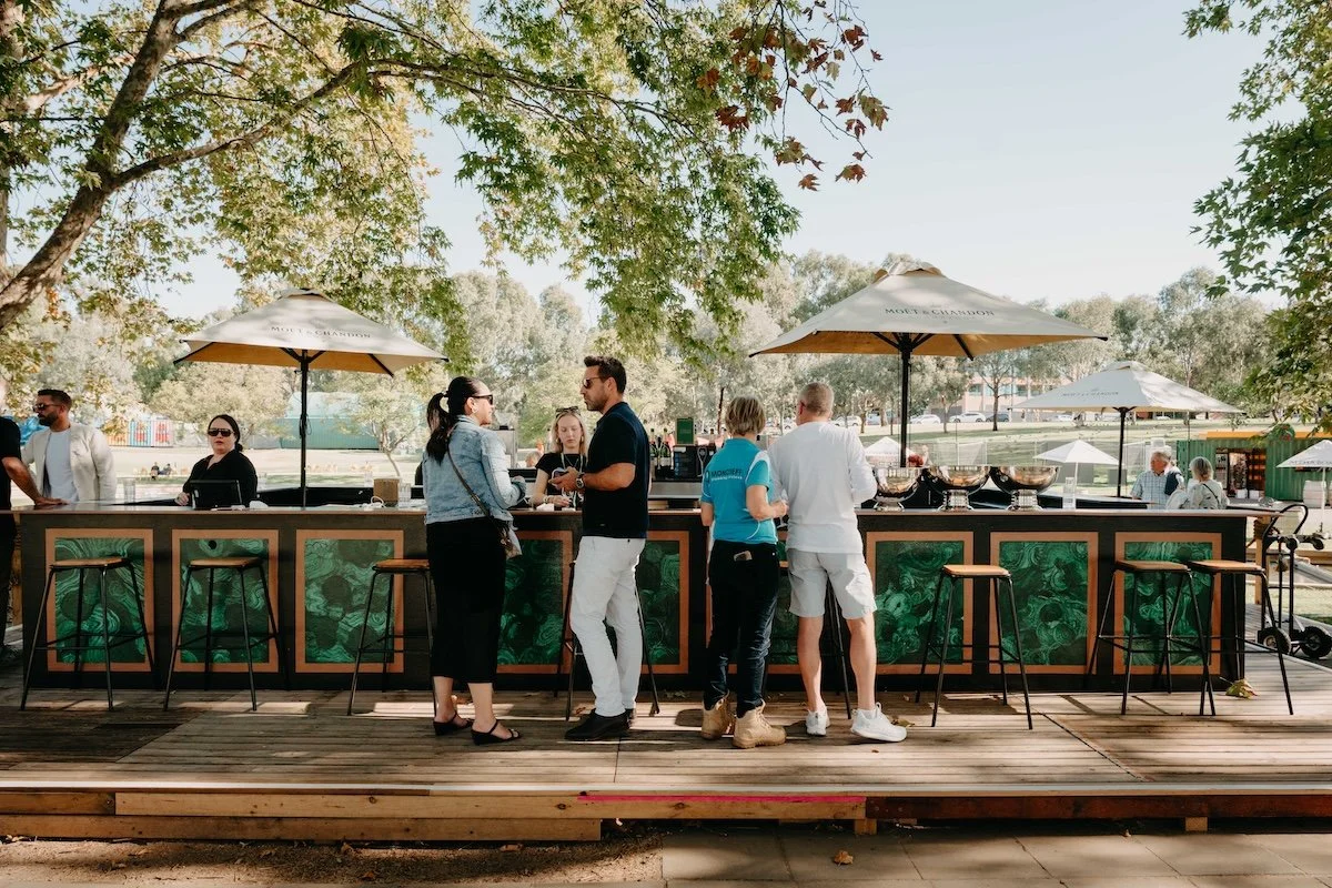 Outdoor bar at Champagne Island in Gluttony, Adelaide, featuring Moët & Chandon umbrellas, a green-paneled counter, and patrons in a sunny park setting.