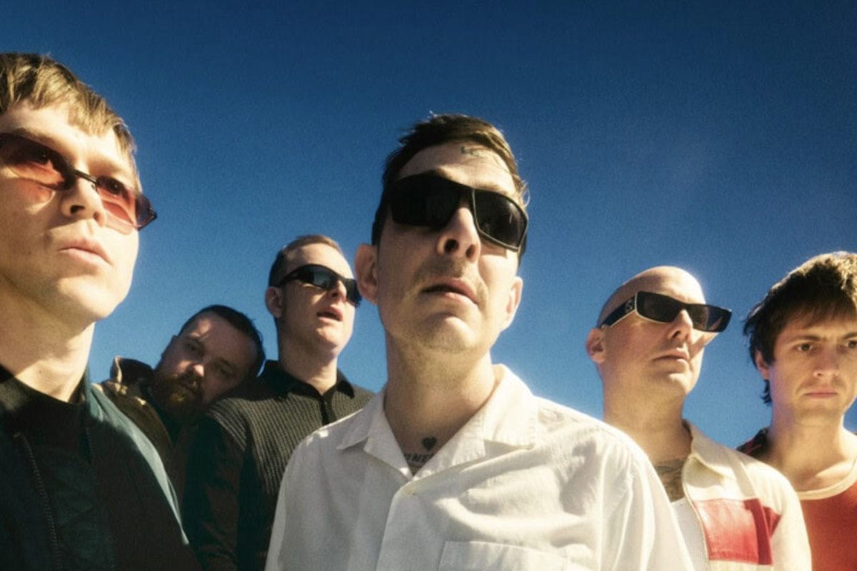 Low-angle shot of the band Viagra Boys wearing sunglasses against a clear blue sky. The lead singer in a white shirt stands front and center.