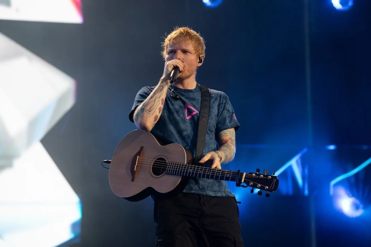 Ed Sheeran performs live on stage, singing into a microphone and playing his acoustic guitar, wearing a dark blue t-shirt and cargo shorts.