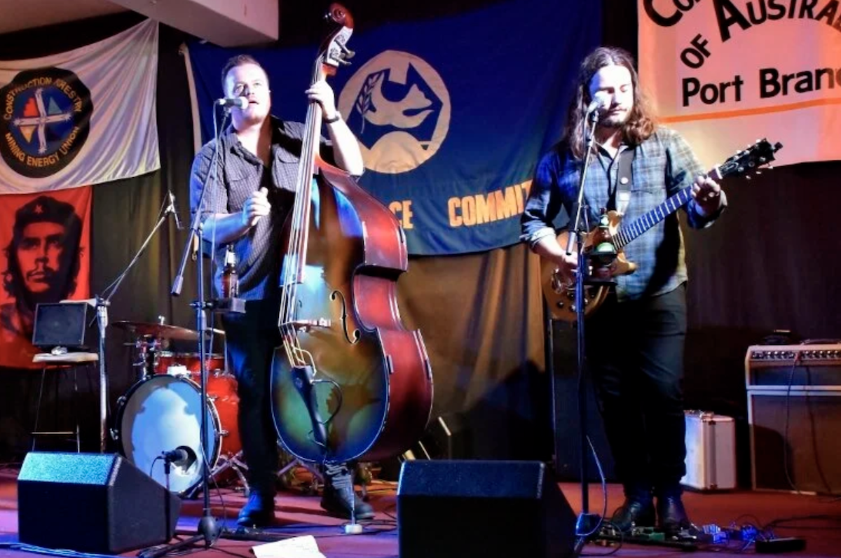 Live band performing on stage at Semaphore Workers Club with a double bass player, guitarist, and union banners hanging in the background.