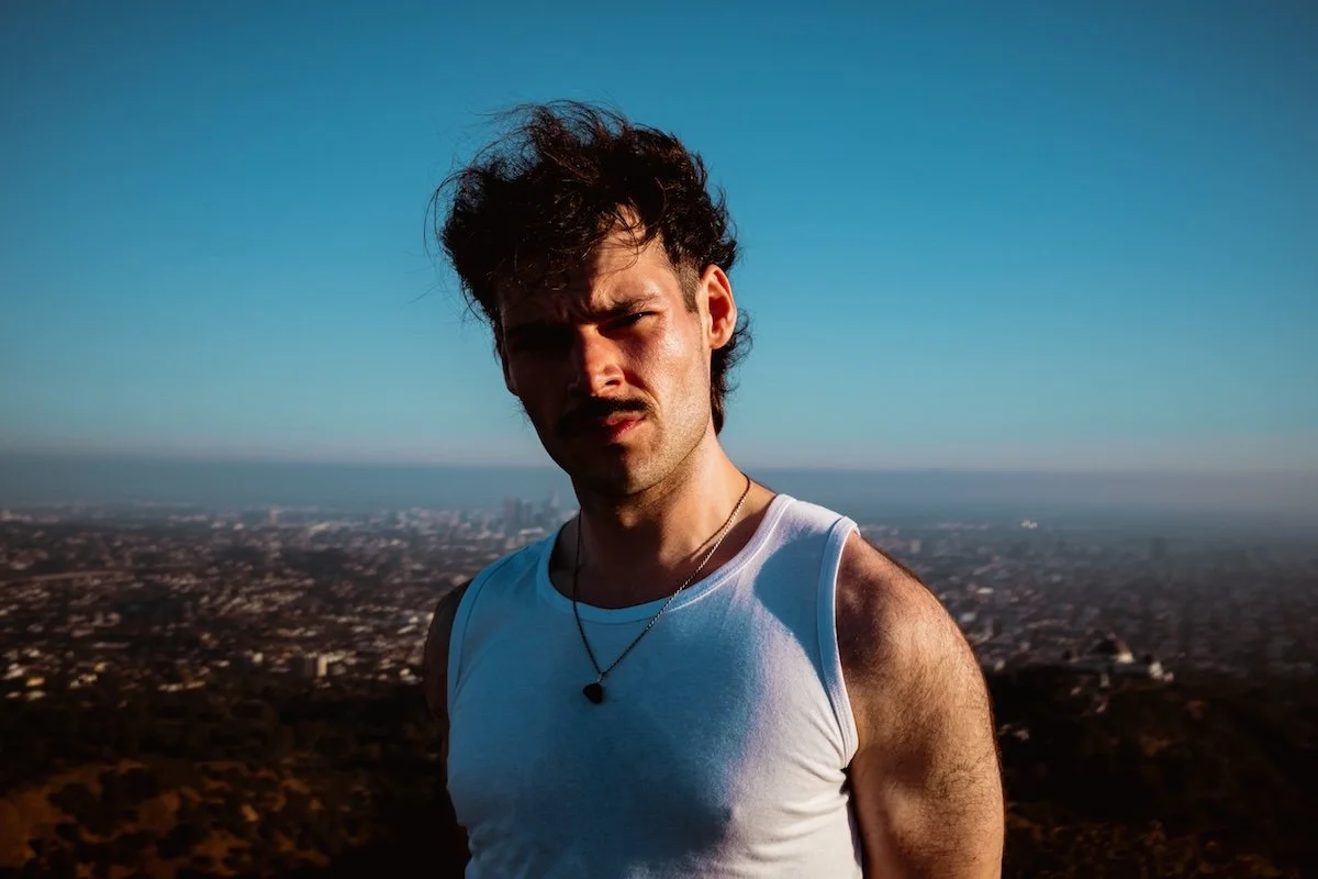 Indie musician James Marriott, wearing a white singlet and mustache, poses on a hill overlooking a sprawling city under a blue sky.