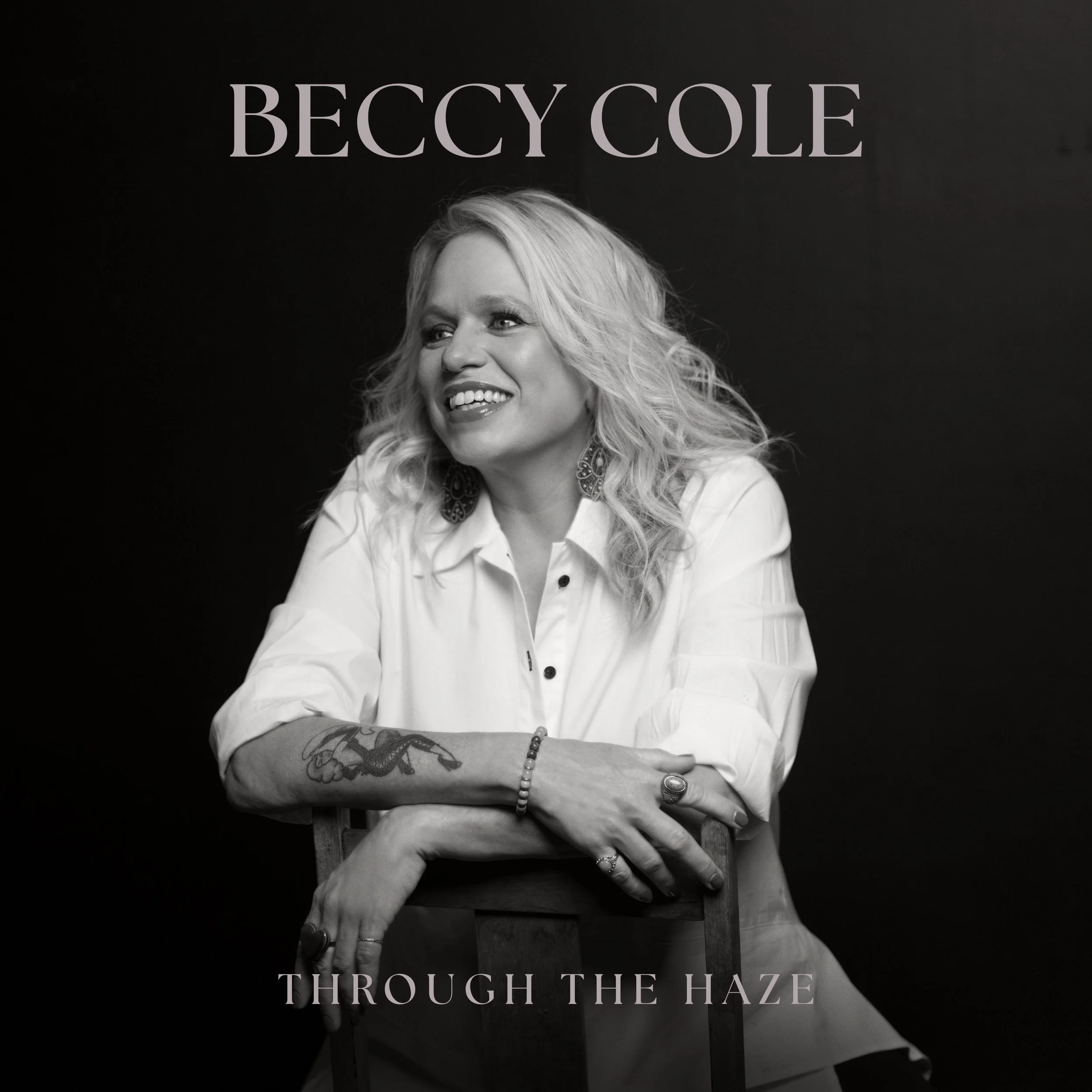 Beccy Cole Through the Haze album cover. Black and white portrait of a smiling Beccy Cole leaning on a chair against a dark, minimalist background.