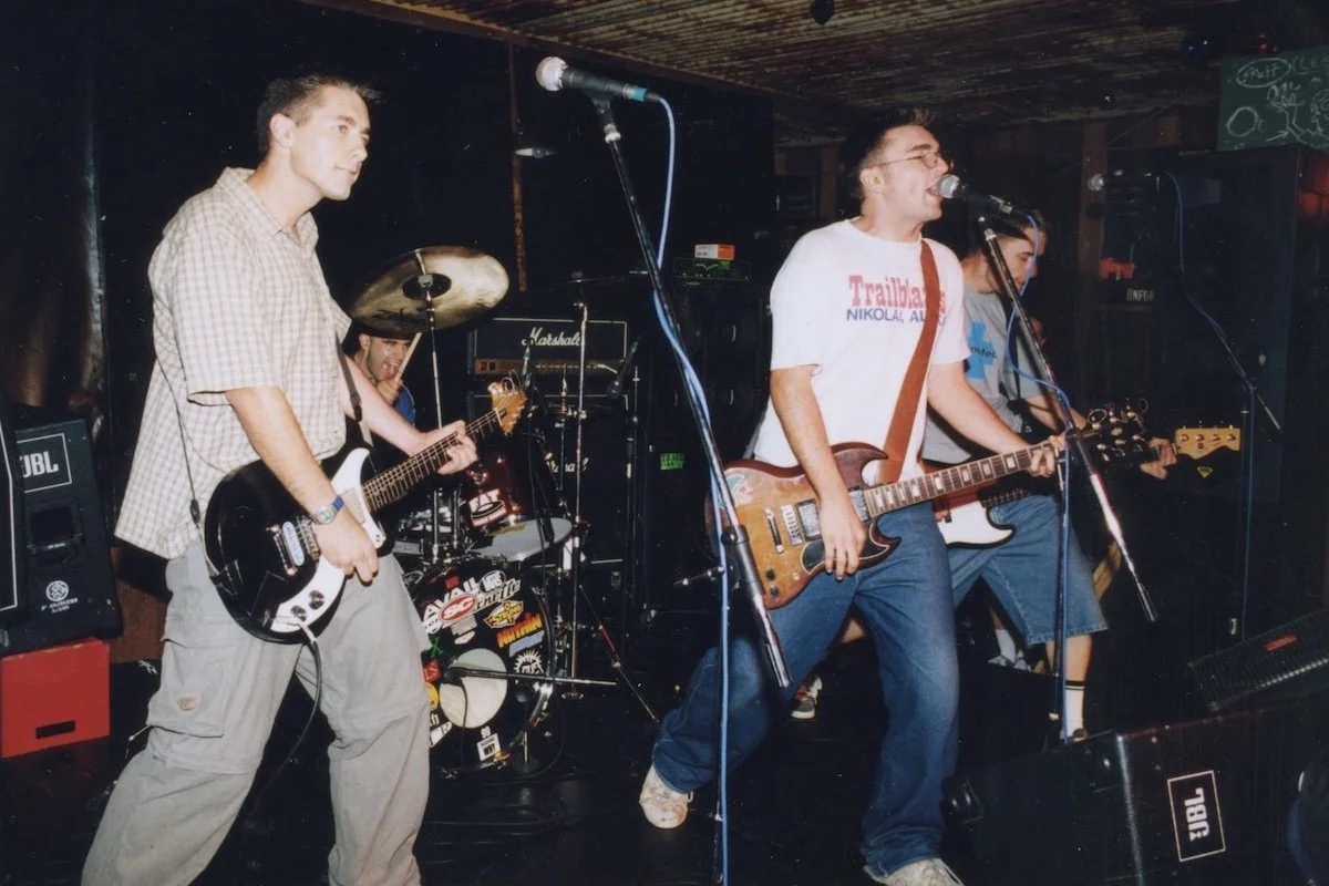 The punk rock band Thinktank performing live on a dark stage. Guitarists are at the front; the singer wears a white shirt, playing a Gibson SG guitar.