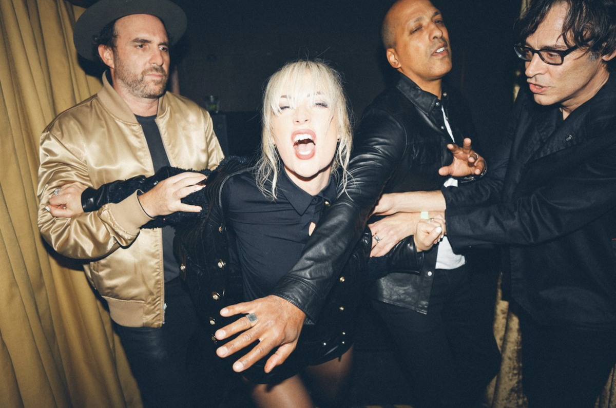 Indie rock band Metric posing in a dynamic, candid shot. Lead singer Emily Haines shouts mid-action, surrounded by bandmates in a dark, moody setting.