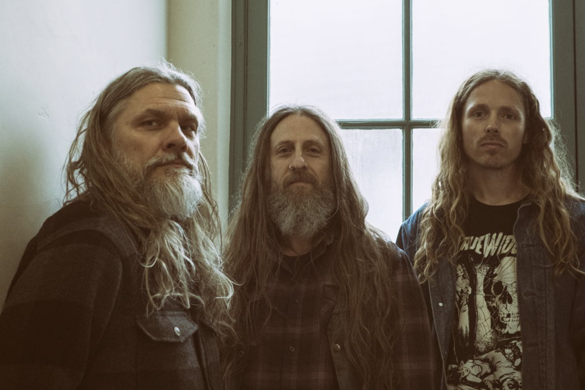 YOB band photo featuring Mike Scheidt, Aaron Rieseberg, and Travis Foster with long hair and beards, standing in front of a window.