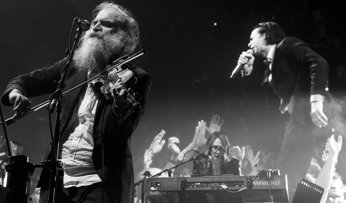 A black and white concert photo of Nick Cave & the Bad Seeds. Warren Ellis plays the violin while Nick Cave sings into the microphone on stage.