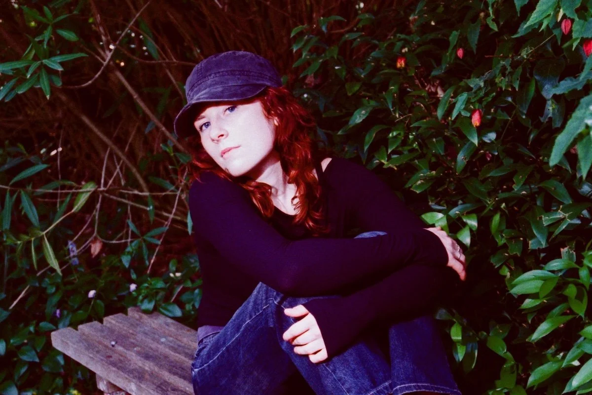 Melbourne indie-pop artist Lucky sitting on a wooden bench outdoors. She has red hair and wears a dark cap and long-sleeved shirt against green foliage.