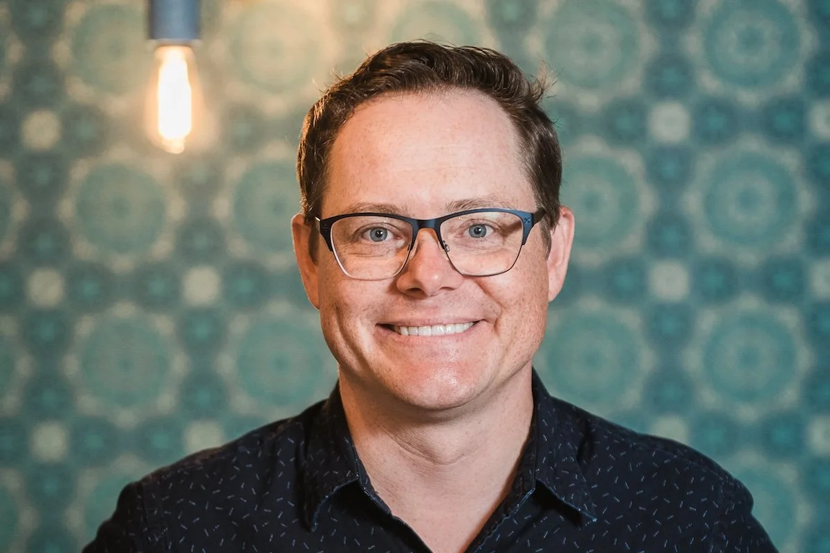 A headshot of Marc Carnes smiling with glasses in a dark patterned shirt, set against a teal mosaic background with a warm light.