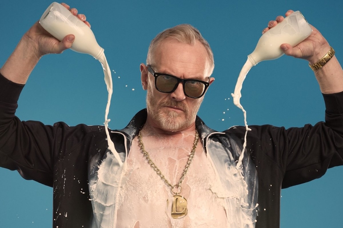 Comedian Greg Davies pours milk over himself in a humorous portrait. He wears sunglasses and a gold chain against a bright blue background.