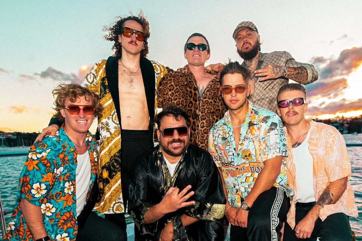 Group photo of 3% and The Terrys posing together outdoors on a boat at sunset, wearing sunglasses, patterned shirts, and robes.
