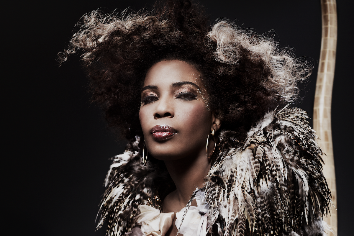 Macy Gray portrait by Giuliano Bekor featuring her signature afro, gold glitter makeup, hoop earrings, and a dramatic feathered outfit.