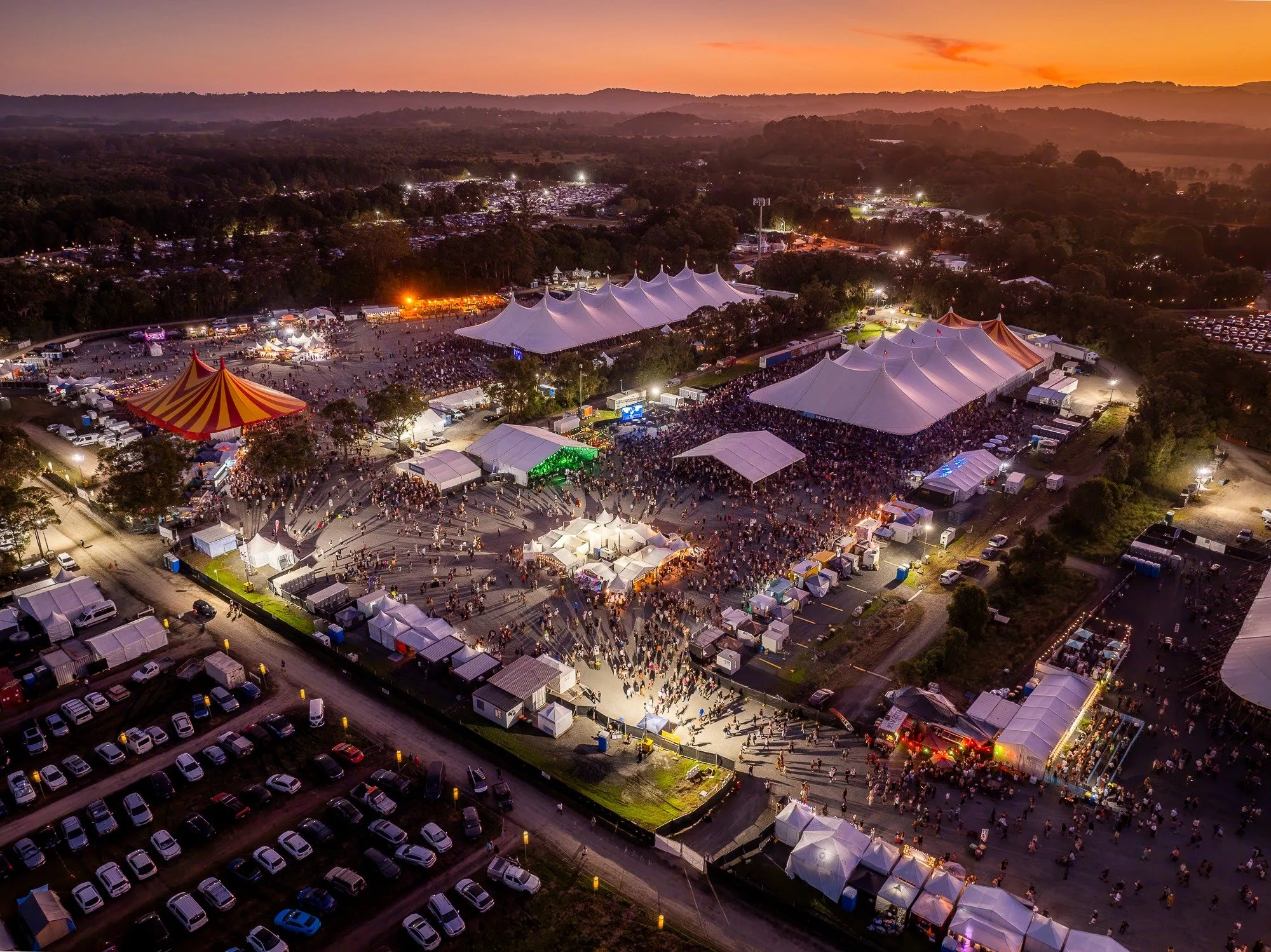 Aerial view of Bluesfest Byron Bay at sunset, featuring large festival tents, crowds, and a vibrant orange sky over the Tyagarah Tea Tree Farm.