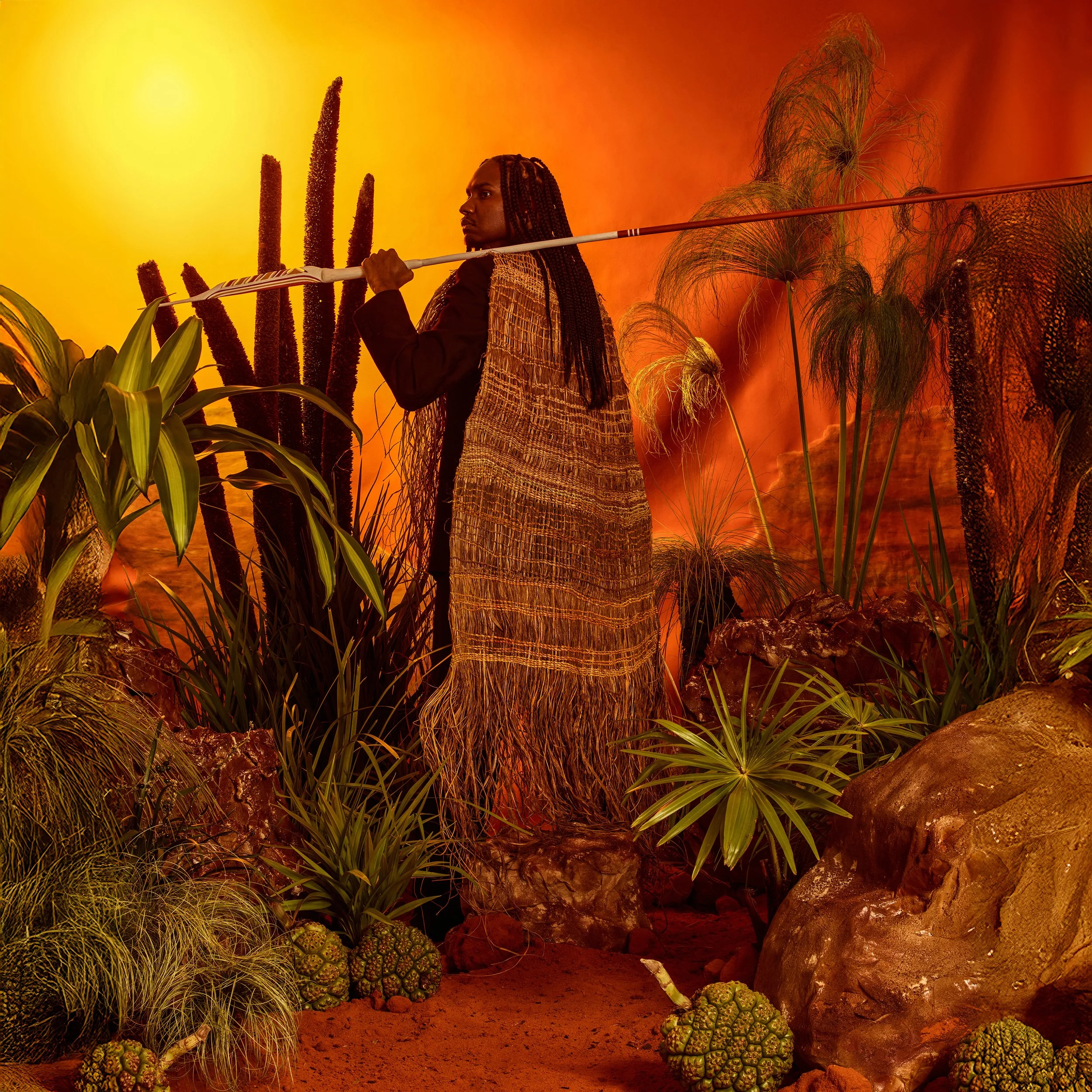 Baker Boy stands in profile wearing a woven cloak and holding a spear, surrounded by lush plants under a warm, glowing sun.