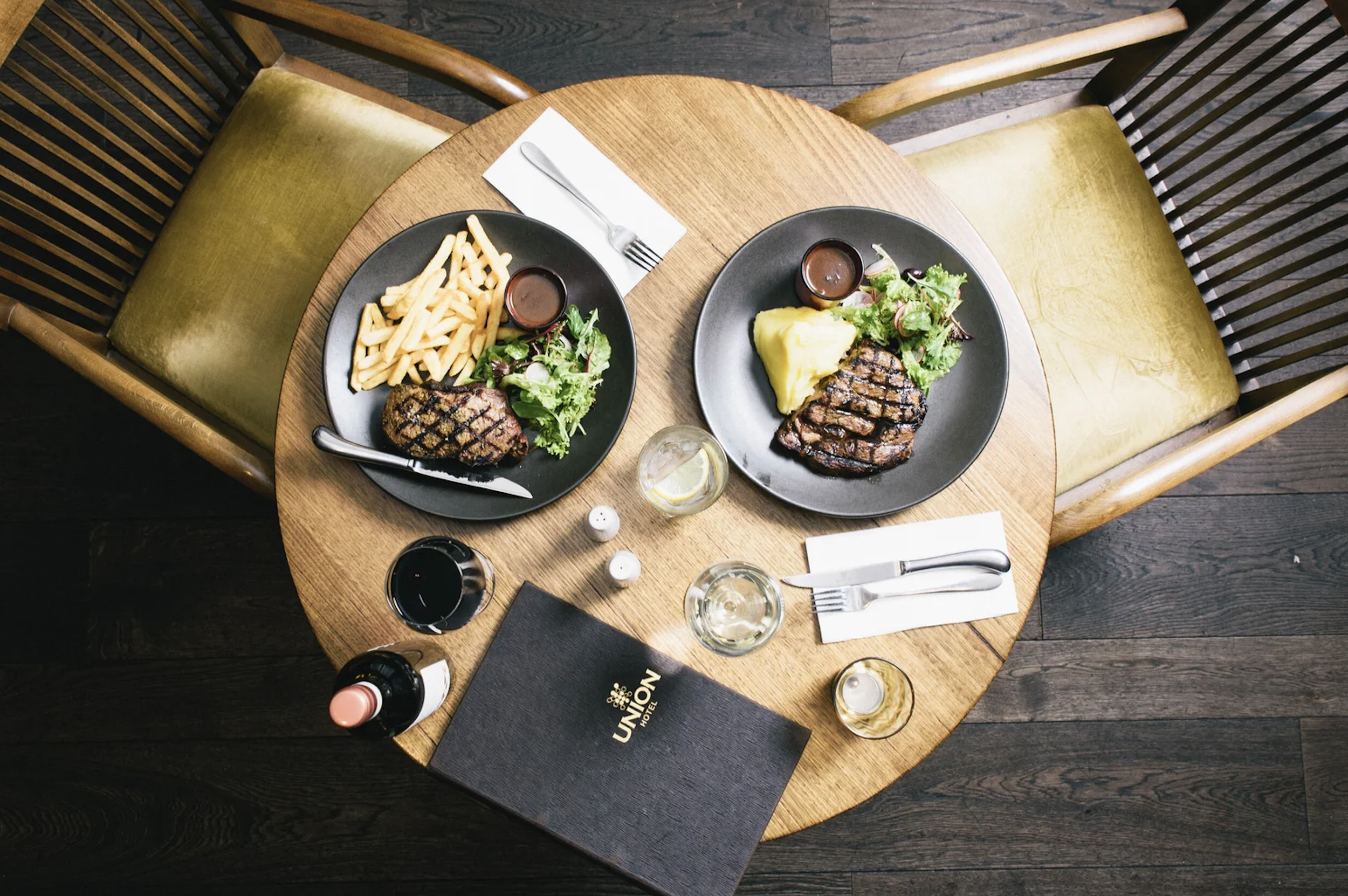 Top-down view of pub meals at Union Hotel Adelaide, featuring grilled steaks with chips, mashed potatoes, salad, wine, and a menu on a wooden table.