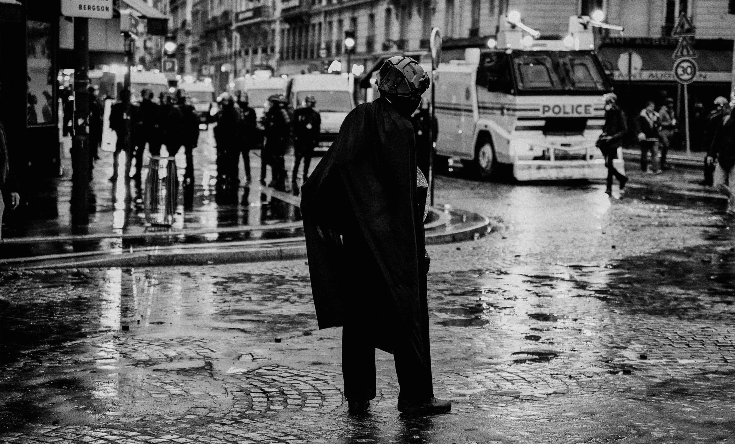 Black and white photograph of a yyoung male body facing a line of police shields, from the series FRICTION by Lulu La Falaise.
