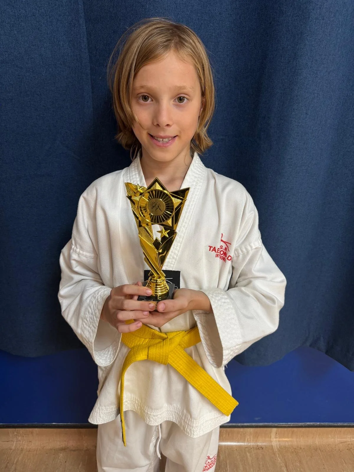 Young girl in martial arts uniform holding a trophy, standing in front of a blue background.