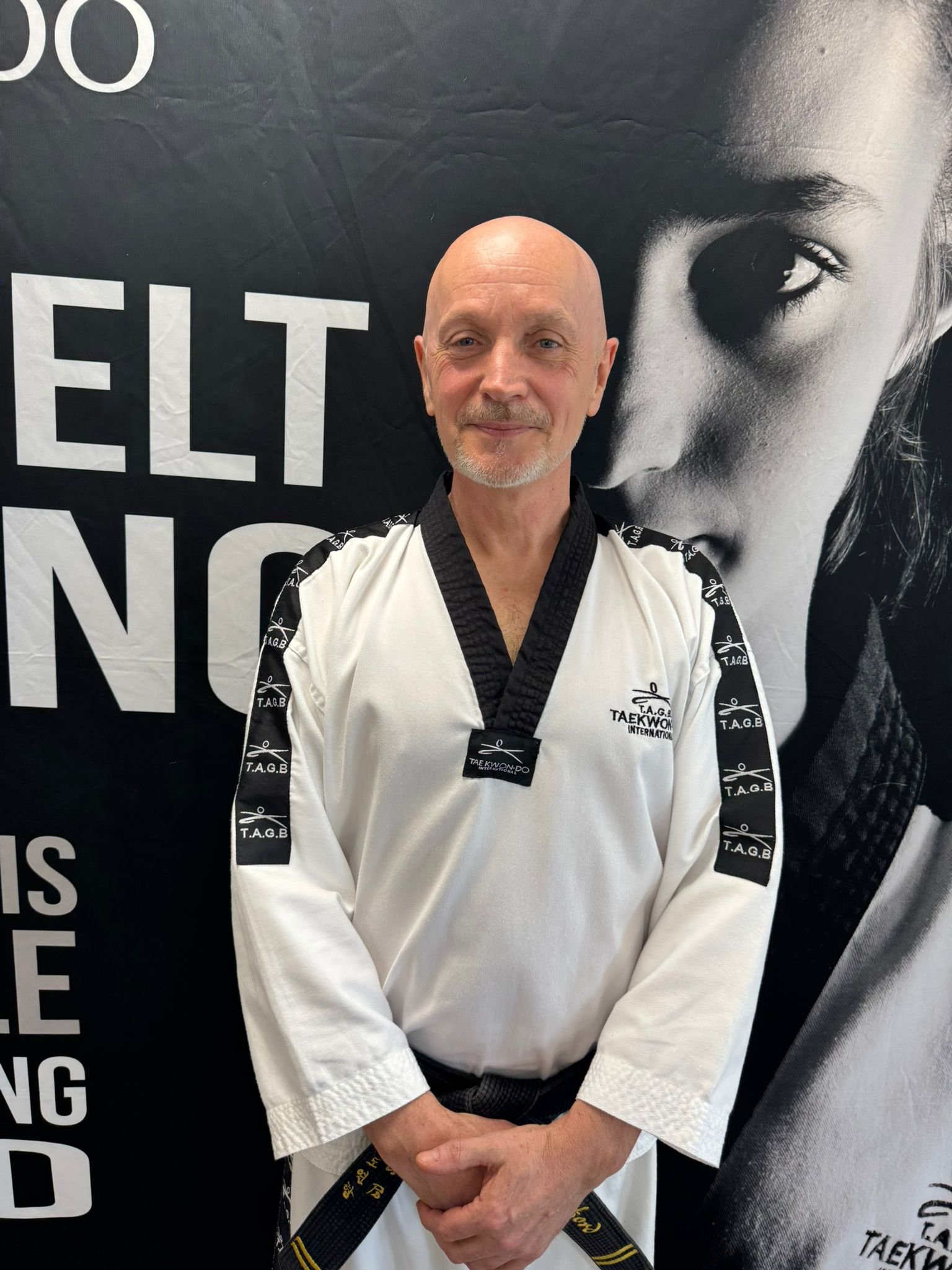Bald man in a taekwondo gi with black belt standing in front of a large black-and-white poster of a woman's face.