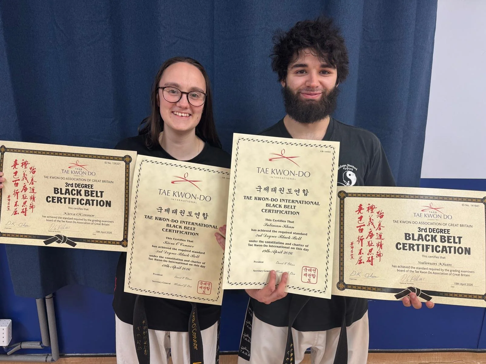 Two individuals, a woman and a man, smiling and holding martial arts black belt certificates. They are dressed in martial arts uniforms and standing against a blue backdrop.