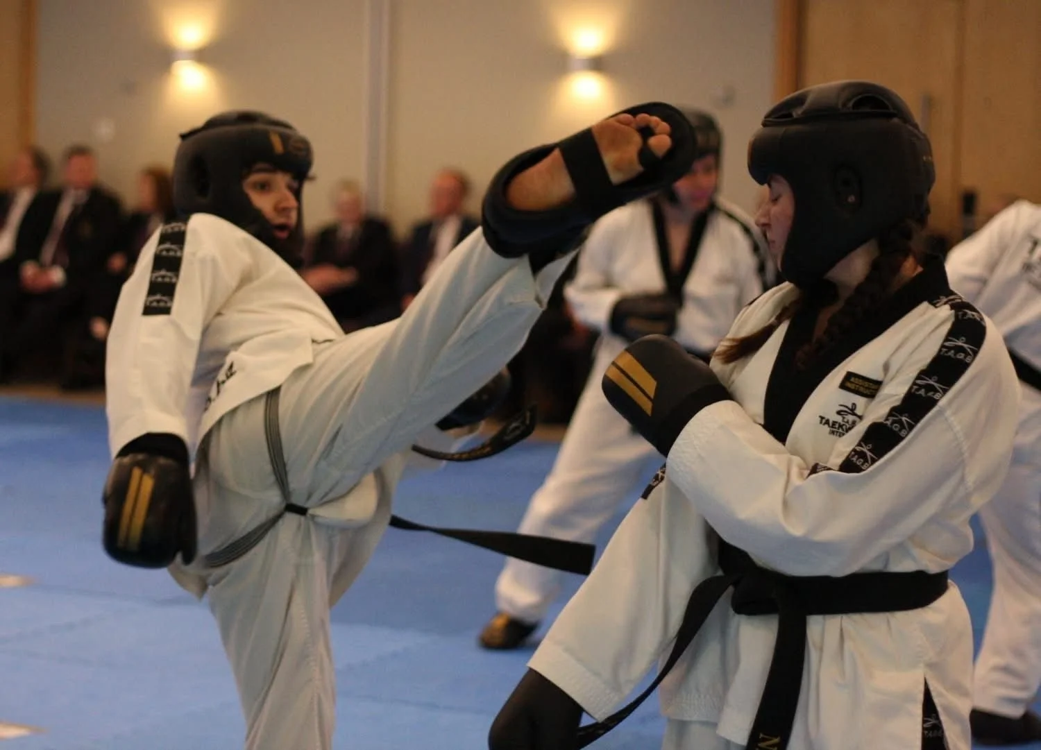 Two women practicing taekwondo sparring, one is executing a high kick, while the other blocks. Both wear white uniforms and black protective gear, including helmets and gloves, in a martial arts competition or class with seated spectators in the back