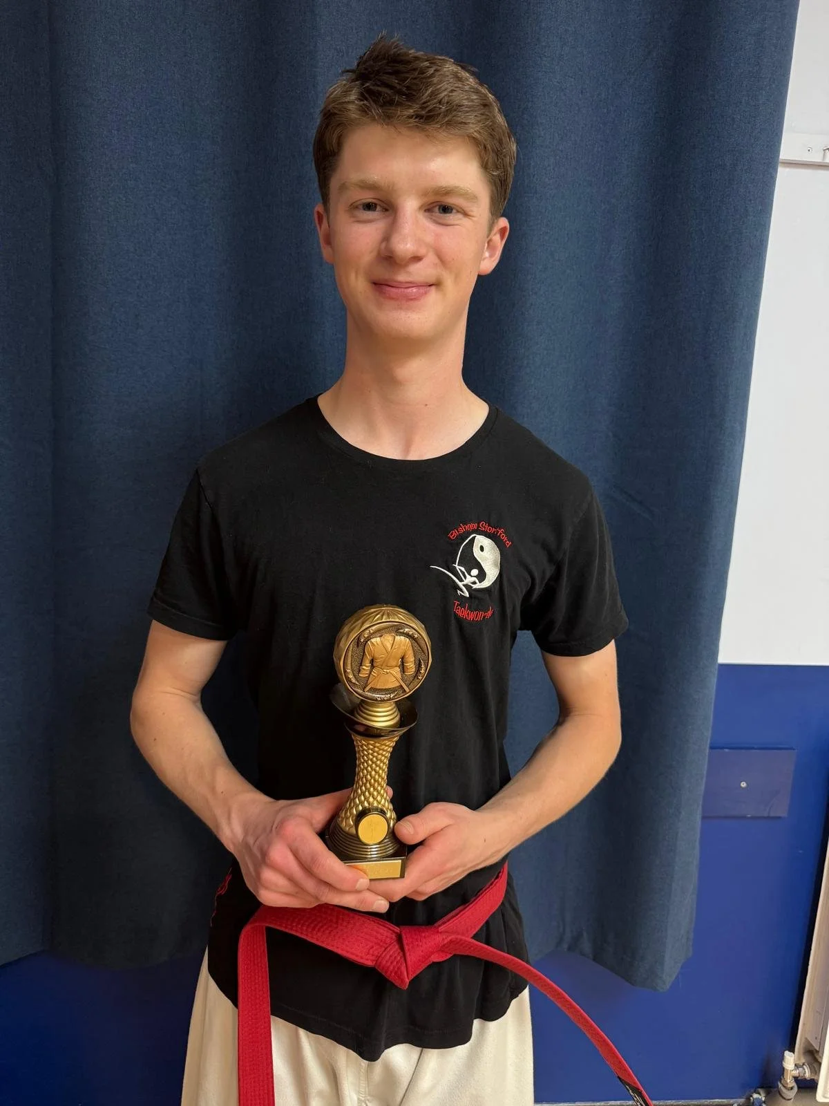 A young man in a black martial arts uniform with a red belt is standing in front of a blue curtain. He is holding a gold-colored martial arts trophy with a karate figure on top and a medallion engraved with a karate kata. He has short, light brown ha