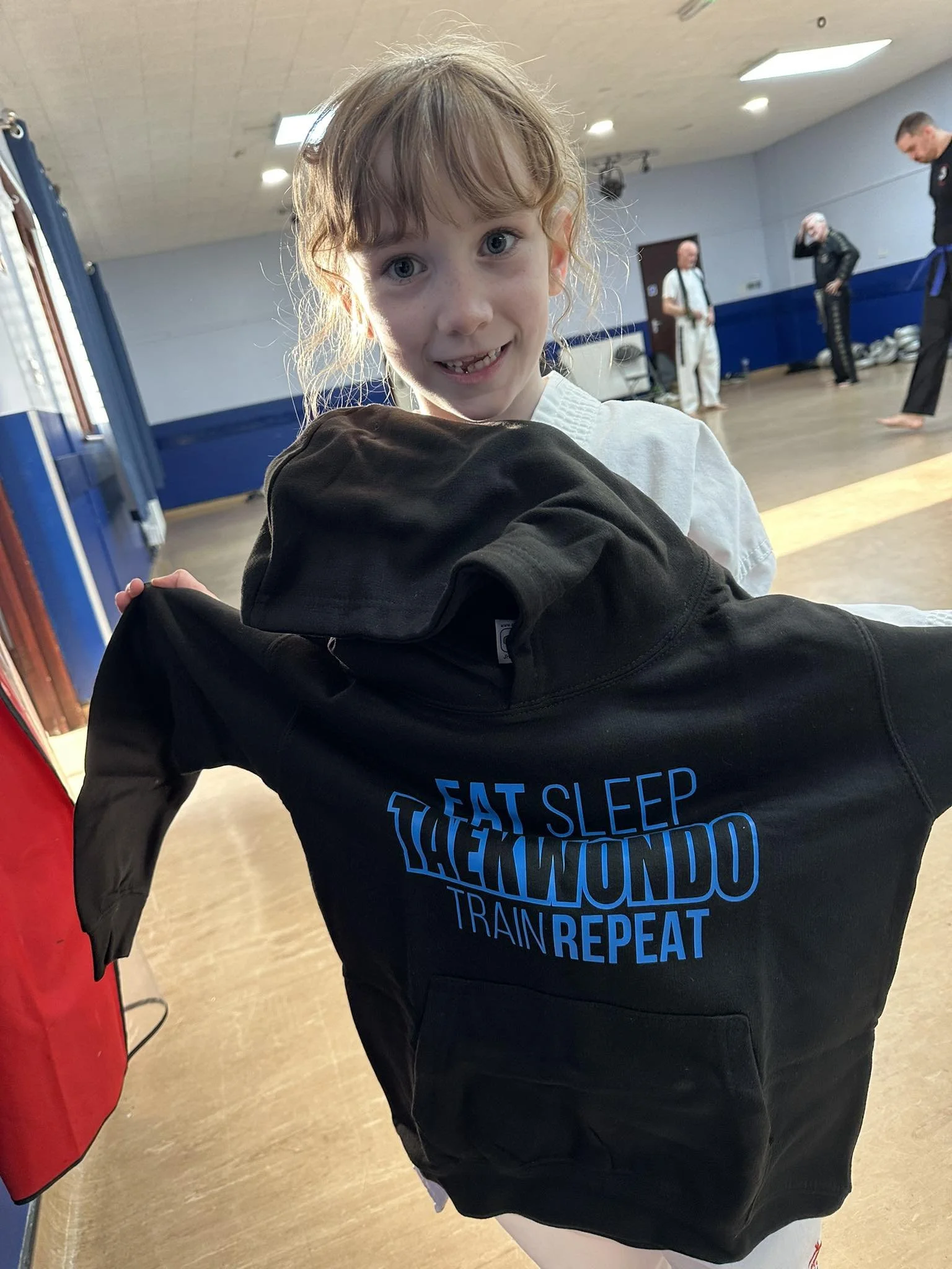 Young girl in a martial arts uniform holding a black jacket with the text 'Eat Sleep JiuJitsu Train Repeat' in a gym, with martial arts instructors in the background.
